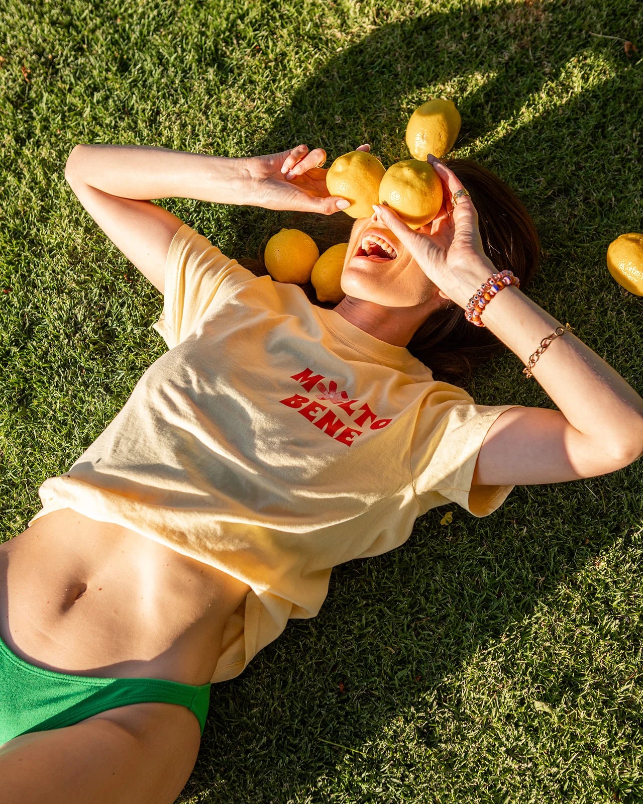 Lounging on grass, a person playfully holds lemons over their eyes while wearing a yellow "Molto Bene" T-shirt by MAKU THE LABEL and green bottoms. Surrounded by hand-painted designs and scattered lemons, their 100% cotton tee shines in the bright sun.