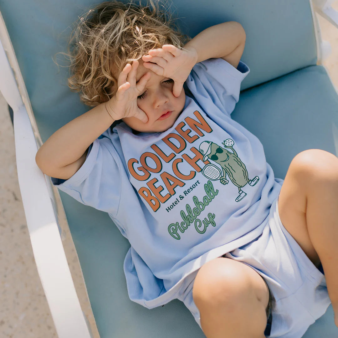 A young child with curly blonde hair is lounging on a chair, dressed in a relaxed-fit Golden Beach Mid Sleeve Tee in Palm Blue and shorts. The tee features "Golden Beach Hotel & Resort Pickleball Cup." The child shades their eyes with their hands, enjoying the sunlight.