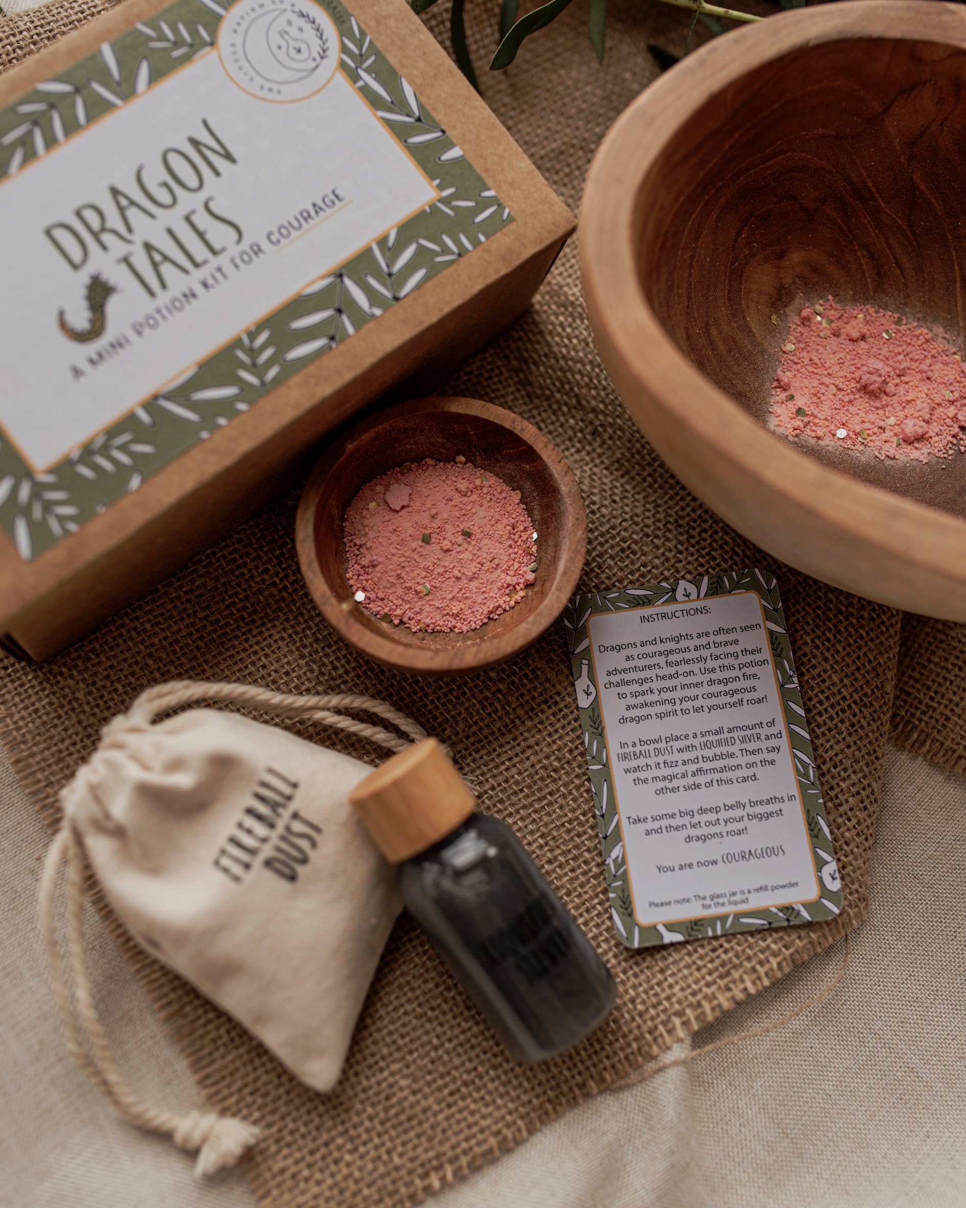 A neatly arranged display features the MINI Dragon Tales Potion Kit by THE LITTLE POTION CO, perfect for imaginative play and offering a potion for courage. It includes a small wooden bowl with pink powder, a larger empty bowl, and a vial of "Elven Dust." A drawstring pouch and instruction card complete the set, all on a burlap-covered surface.