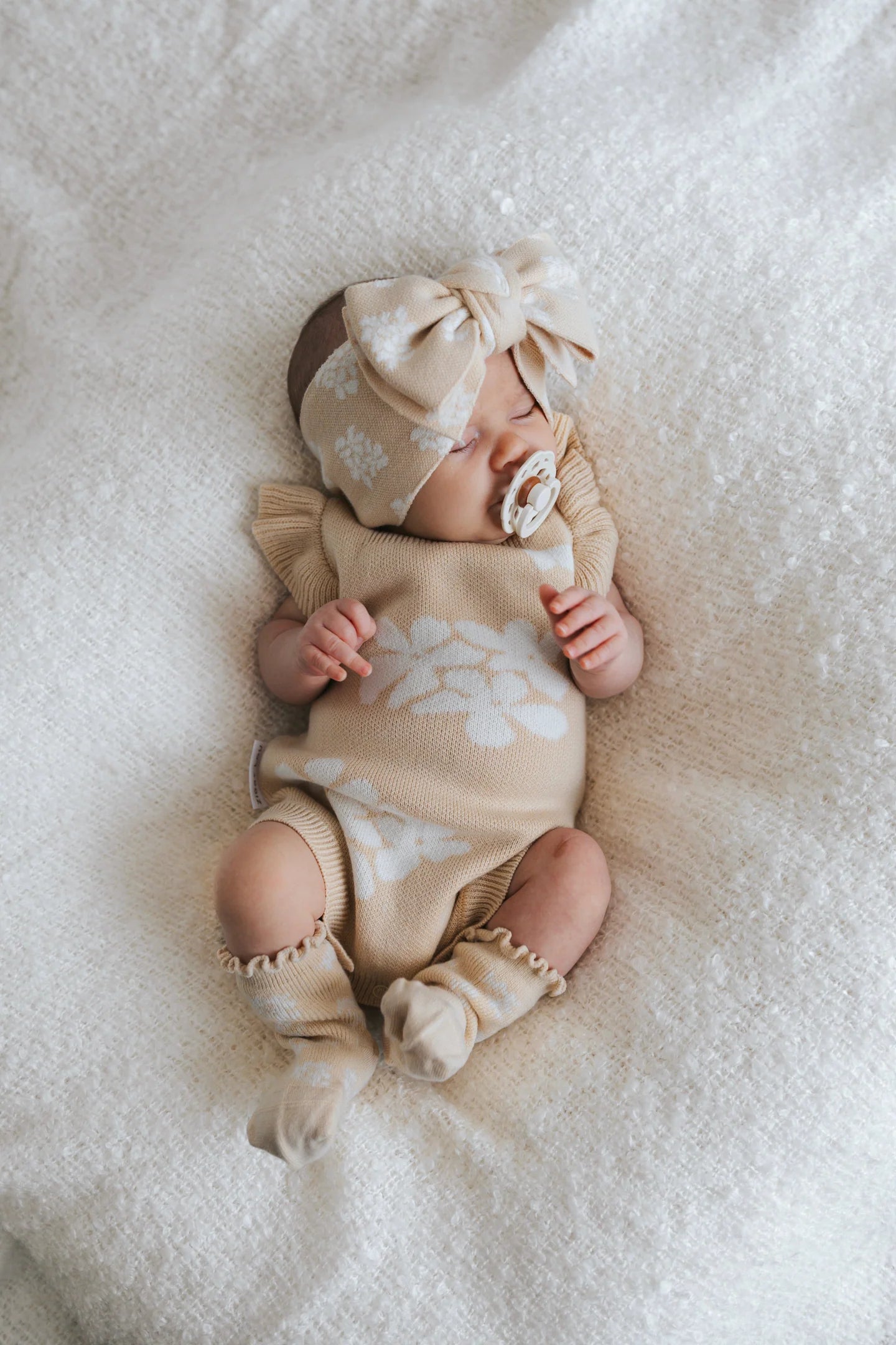 A baby lies on a soft, white blanket, dressed in a beige ZIGGY LOU Frill Romper Banksy adorned with white floral patterns. The infant accessorizes with a matching large bow headband and a pacifier. The ensemble features functional snaps, ruffled sleeves, and coordinating socks.