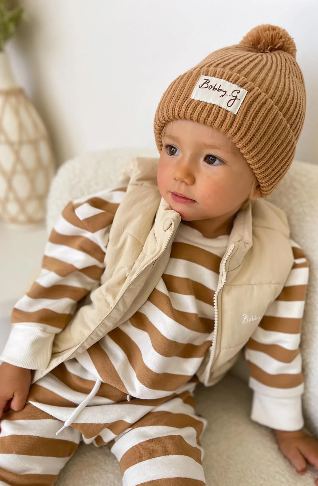 A young toddler wearing the Camden caramel striped pullover and matching pants with a puffer vest over the top an the pom pom beanie in rusty.