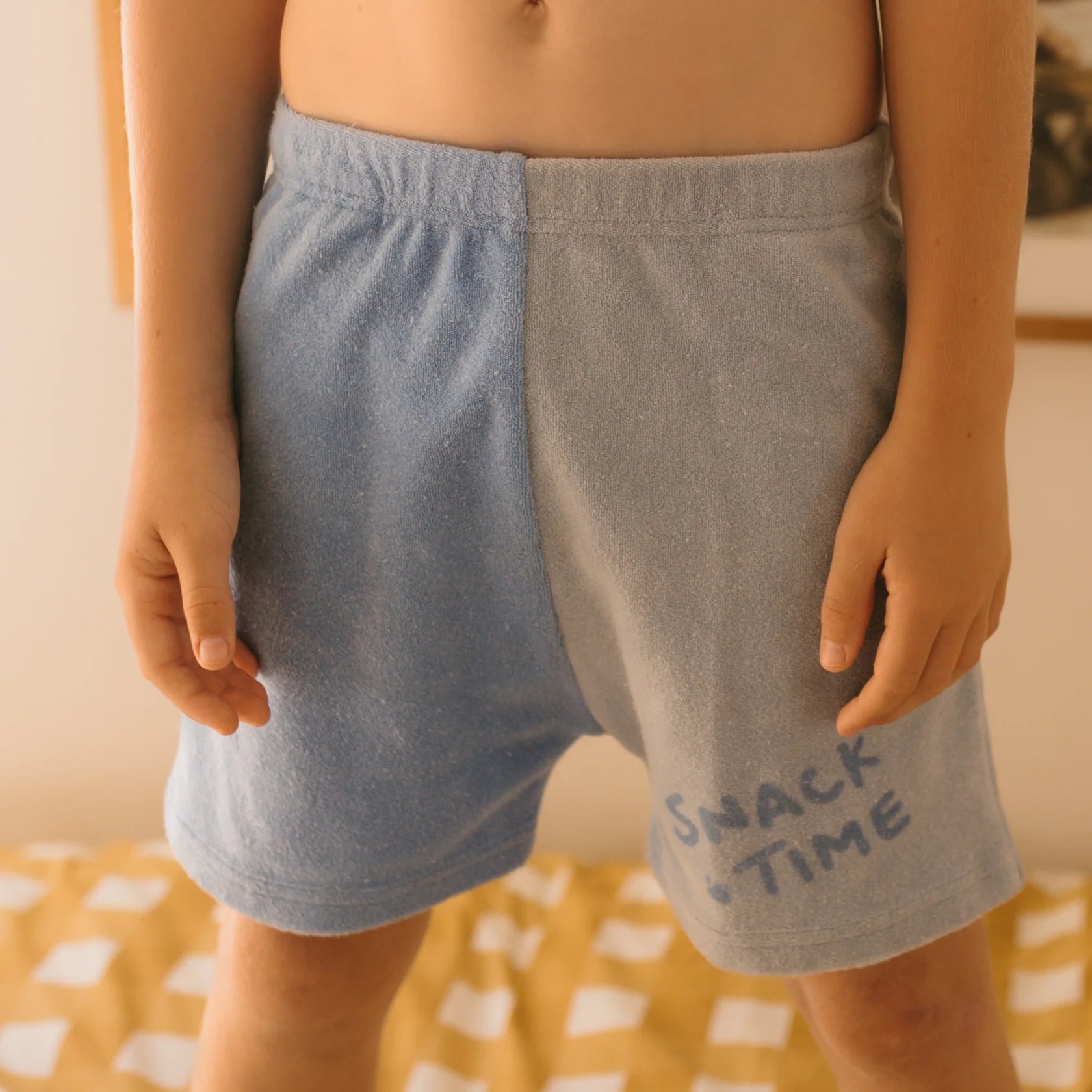 A young child stands wearing the Play Lounge Shorts Pool from GOLDEN CHILDREN, crafted from soft terry towel cotton. The shorts feature a dark blue "SNACK TIME" print on the right leg and are designed in a two-toned style with one half in light blue and the other in a slightly darker blue.