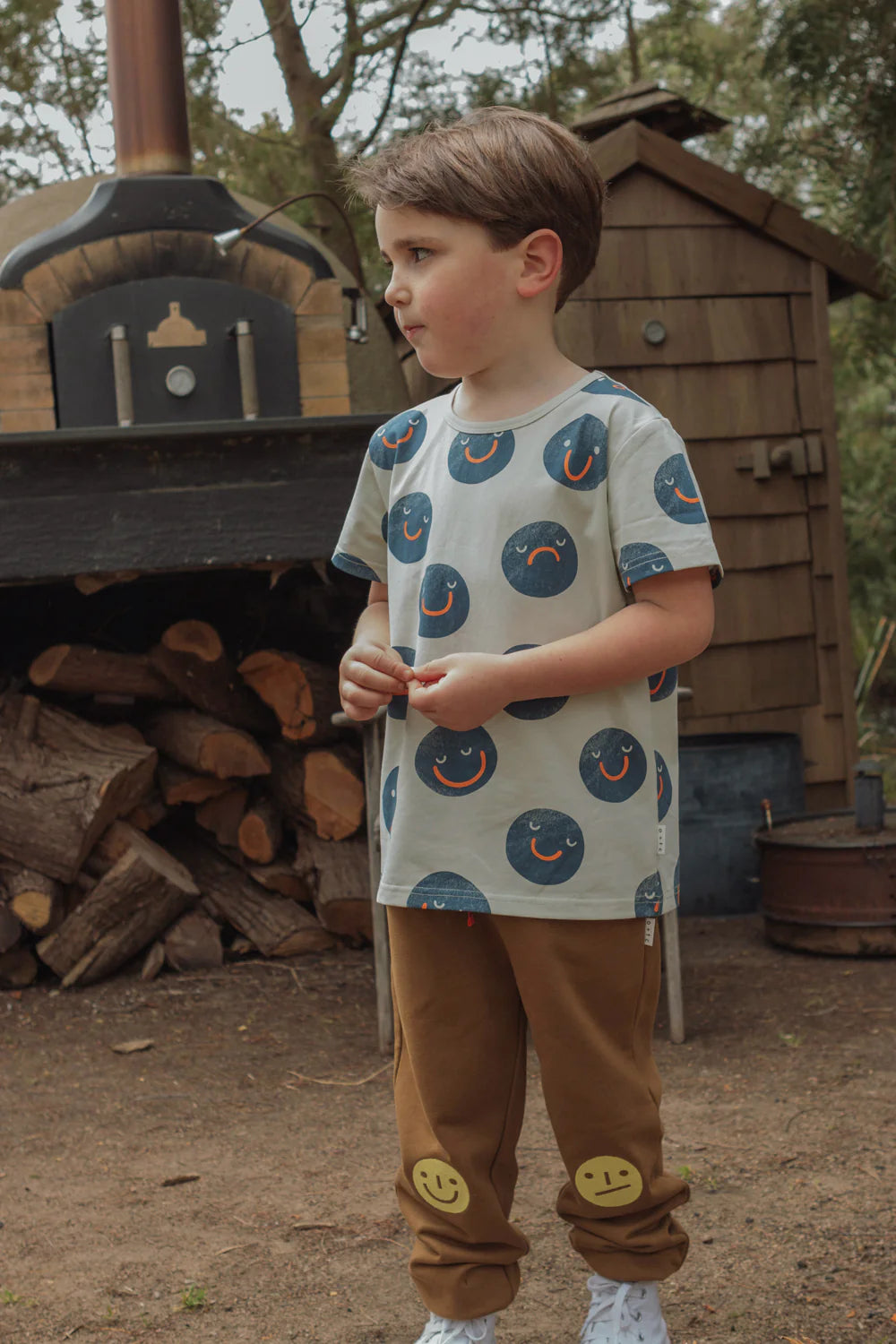 A young child stands outdoors wearing a shirt with fun mood faces and OLIVE + THE CAPTAIN's Mood Track Pants, which feature smiley faces on the knees. Behind the child is a stack of firewood and a wood-fired oven. The background showcases trees and a wooden structure, completing the cozy scene with premium cotton fleece garments.