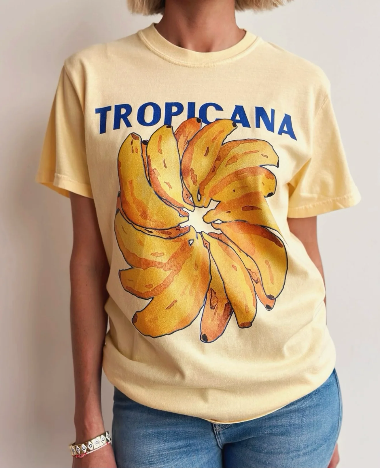 A person wearing a pale yellow Tropicana Banana T-Shirt by MAKU THE LABEL, featuring "TROPICANA" in blue letters and a hand-painted banana design, paired with blue jeans against a light-colored wall.