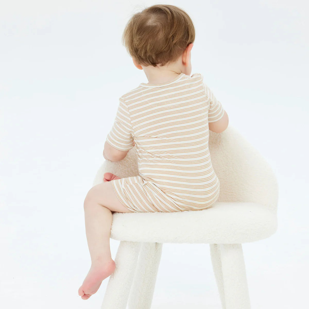 A toddler wearing the Day or Night Short Sleeve Onesie Neutral from KYND BABY, crafted from sustainable viscose fabric, sits on a soft white chair with their back towards the camera. The plain white background provides a serene and gentle setting, perfect for newborn skin.