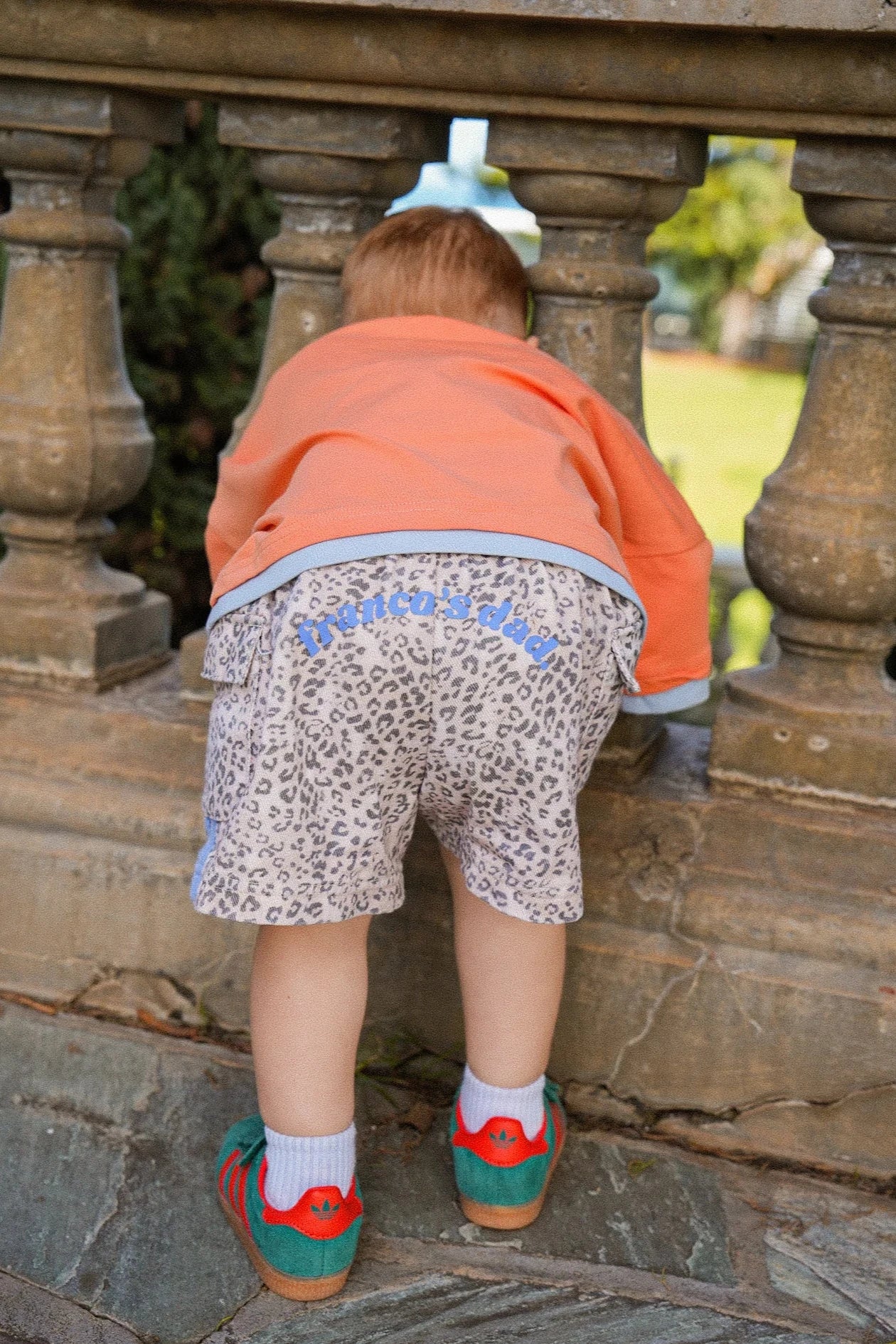 A toddler wearing the PRE-ORDER Leopard Cargo Shorts from FRANCO'S DAD and an orange jacket leans against a stone balustrade, peering through its gaps. The child sports colorful dinosaur shoes, with the greenery in the background adding a fresh touch to the scene.