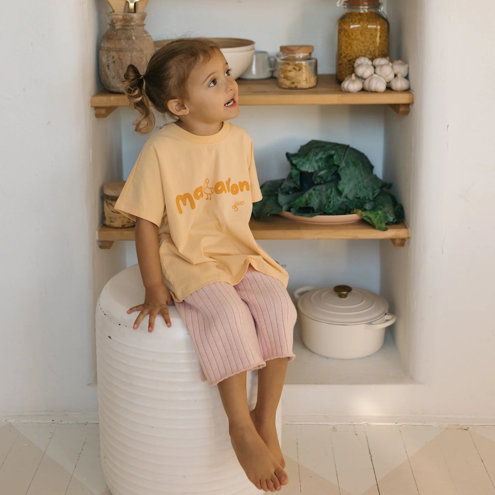 A young child wearing a GOLDEN CHILDREN Macaroni Tee Golden Butter made of soft stretch fabric paired with pink pants is seated on a white stool in a kitchen. Behind them, shelves display jars, a garlic bulb, and leafy greens. The child looks upward with a curious expression.