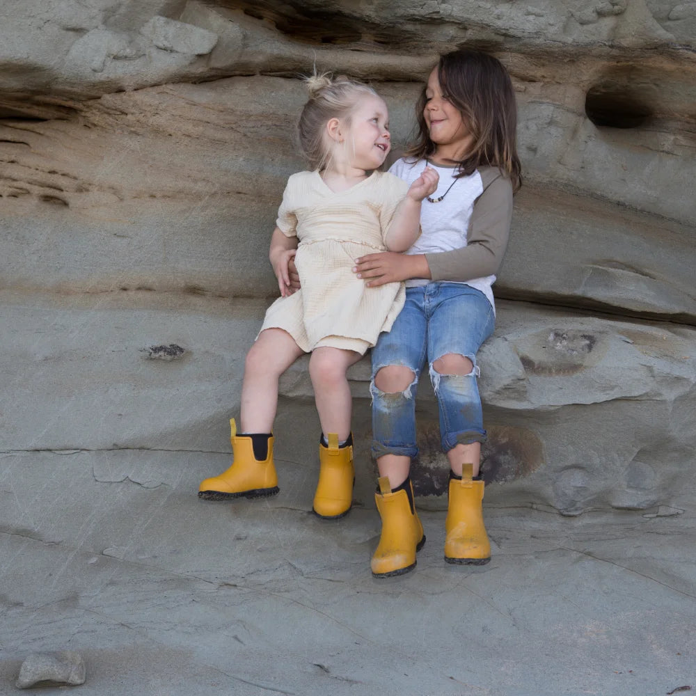 Two children sit on a rock ledge, laughing. The child on the left wears a light outfit and Merry People ~ Bobbi Kids Gumboot Mustard Yellow, while the right sports a brown shirt, ripped jeans, and matching waterproof boots. A textured rock wall stands behind them.