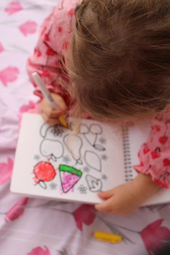 A blonde child in a pink dress showcases creativity using the Oh My Frankie ~ Oh My Scribbles Reusable Colouring Book. The page is full of colorful shapes and hearts, crafted with non-toxic markers, while holding a colored pencil against a pink background.