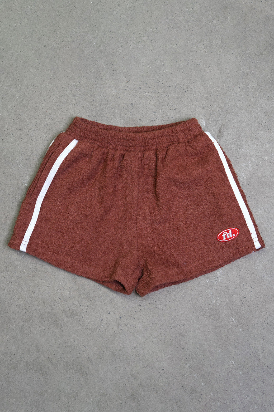 The PRE-ORDER Towel Shorts by FRANCO'S DAD are a summer essential featuring a comfortable elastic waistband and stylish white side stripes. Crafted from lightweight towelling material in brown, they showcase a small red logo patch on the bottom left against a grey background, all in a trendy baggy fit.