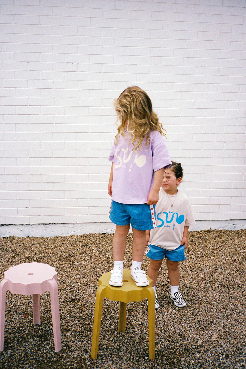 In front of a white brick wall, two children are outdoors; one stands on a yellow stool wearing an oversized Sunday Siblings Il Sole Tee in Purple/Cream. The other, in a white cotton shirt, stands nearby on the gravel. Both wear blue shorts and sneakers with a pink stool beside them.