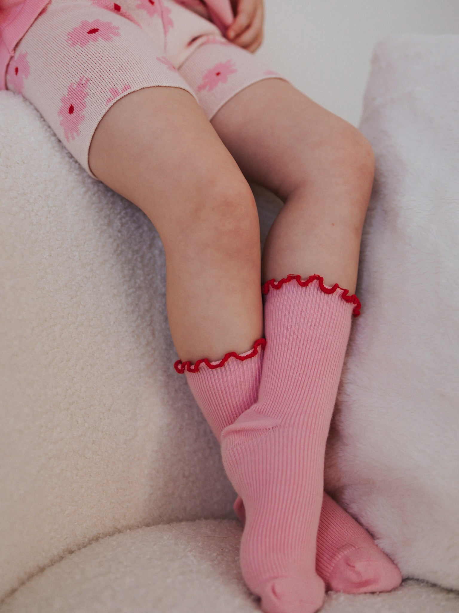 A child sits on a plush white surface, wearing pink shorts with flower patterns and ZIGGY LOU's Ziggy Lou ~ Socks Elodie. These pink knee-high socks with red ruffle trim offer a blend of comfort and style, ideal for mid-lounge moments.