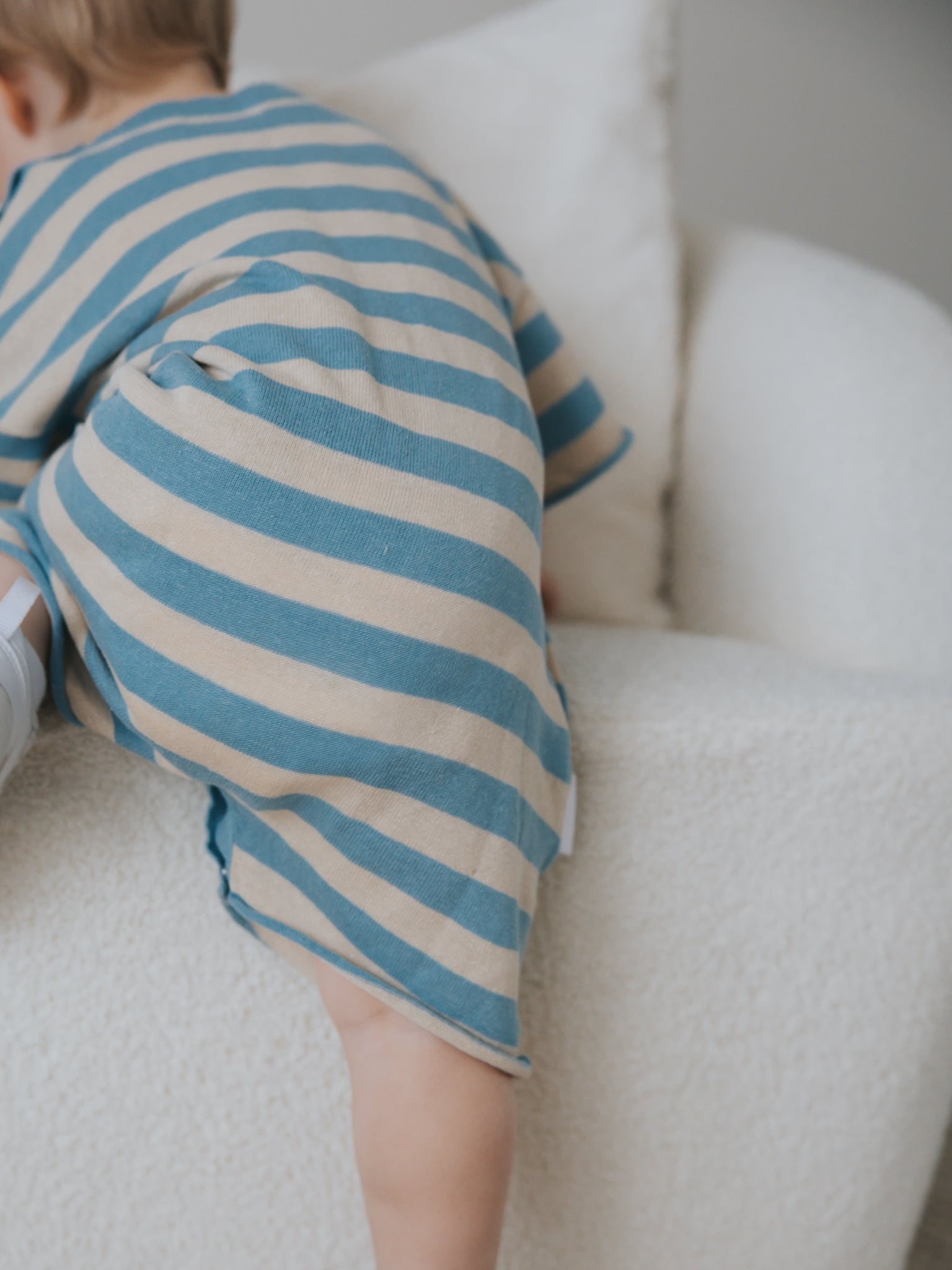 A baby in a Ziggy Lou ~ Playsuit Lake by ZIGGY LOU climbs on a soft, white chair, with only part of their body visible against the chair's textured fabric.