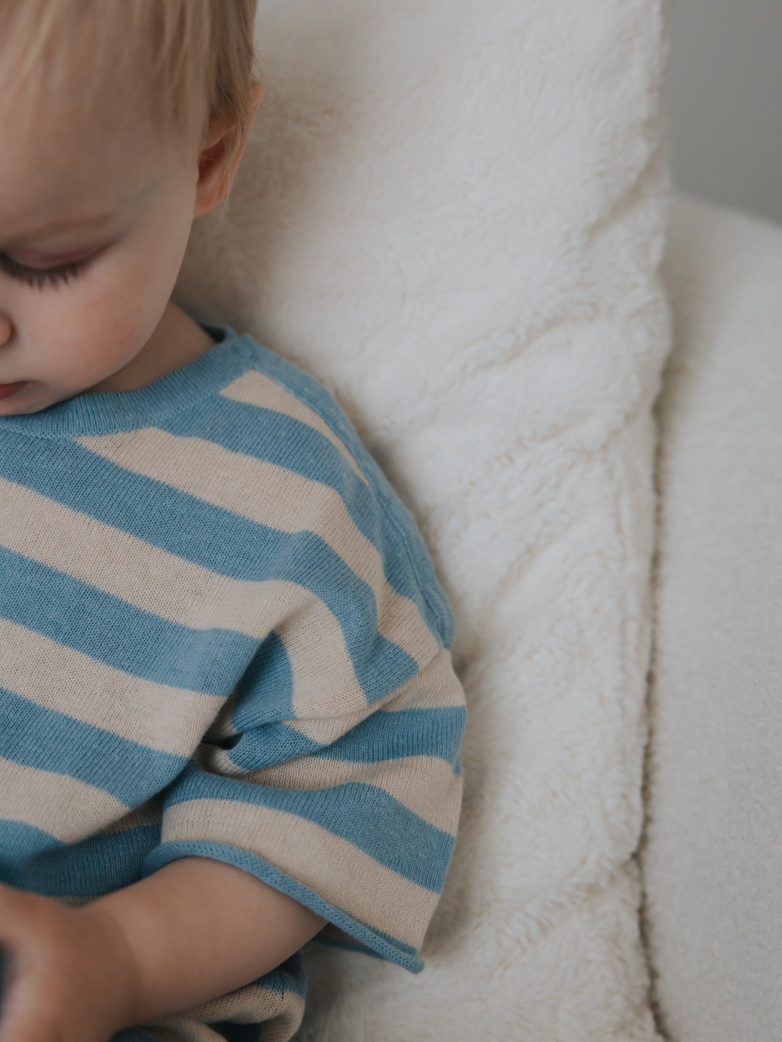 A young child is sitting on a fluffy white cushion, wearing the Ziggy Lou ~ Playsuit Lake from ZIGGY LOU, showcasing its blue and cream stripes. Only part of the child is visible, highlighting the playsuit's pattern and the texture of the cushion.