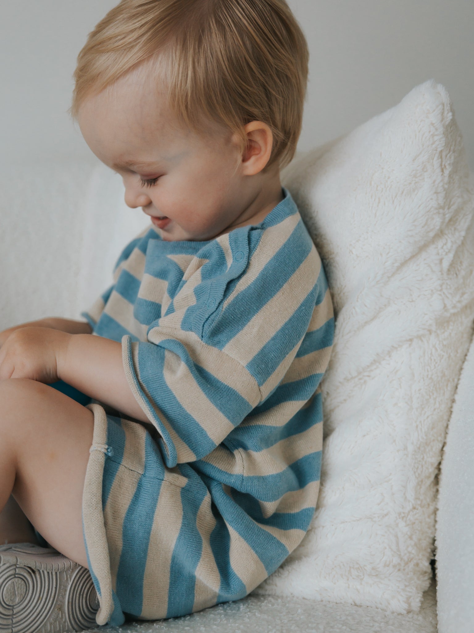 A blonde toddler sits on a soft white couch, wearing a Ziggy Lou playsuit in blue and beige stripes. They focus intently on something in their hands while smiling gently, with a fluffy white pillow behind them.