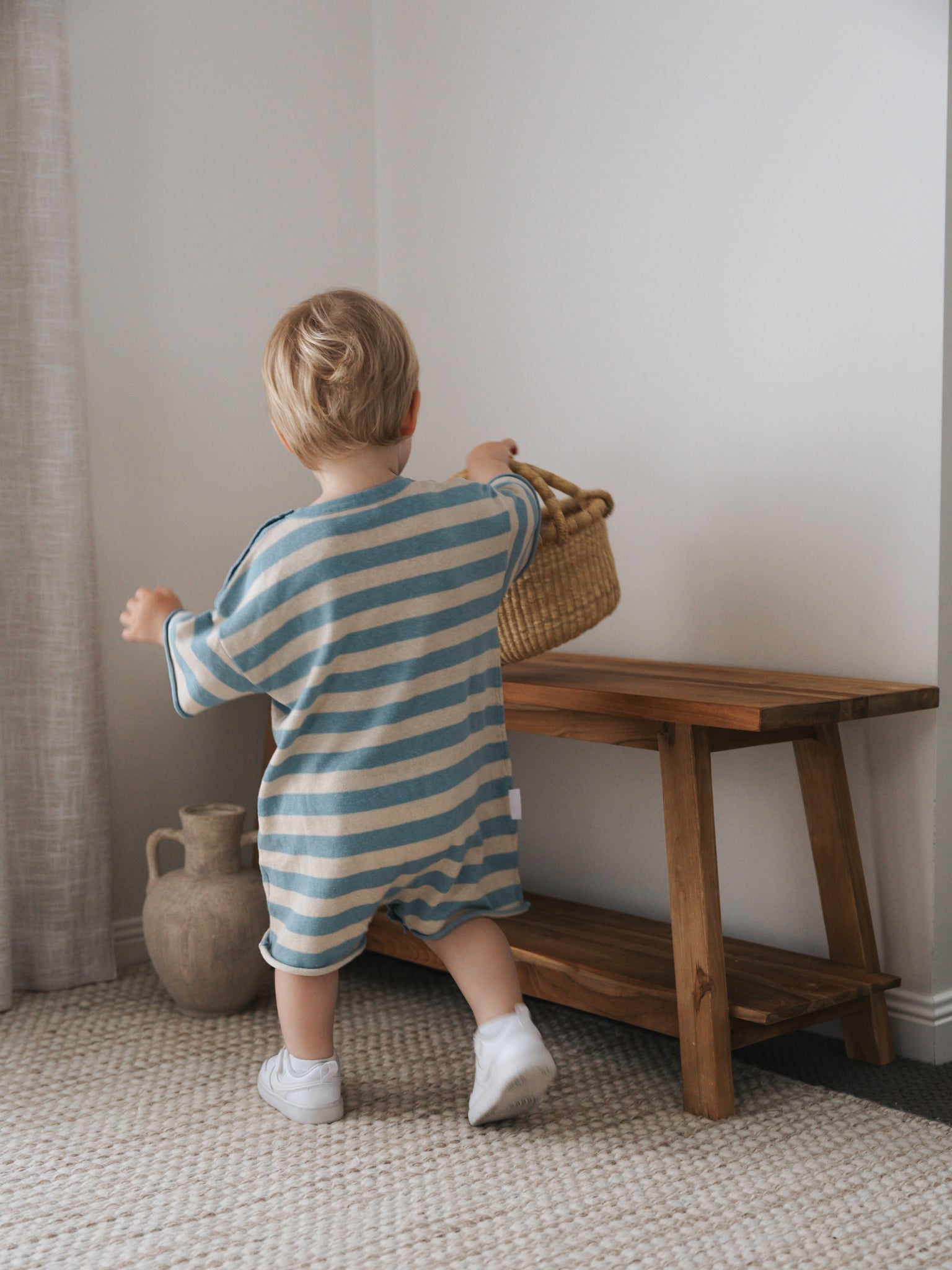 A toddler in a Ziggy Lou ~ Playsuit Lake stands on a carpet, facing away, holding a wicker basket with one foot raised. A wooden bench and ceramic jug enhance the serene vibe of the Costa + Lake Collections against a white wall.