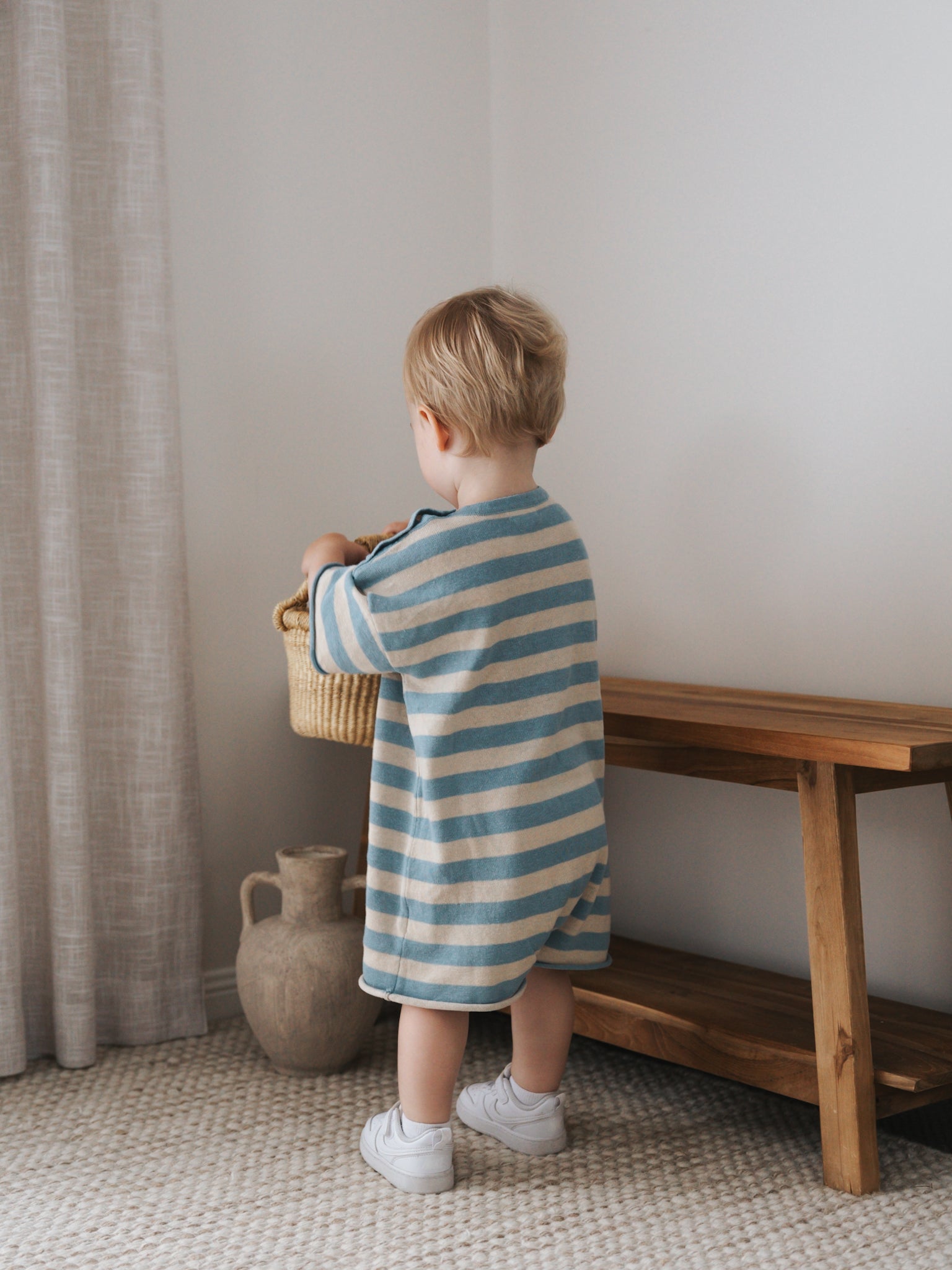 A toddler stands with their back to the camera, wearing a Ziggy Lou ~ Playsuit Lake by ZIGGY LOU. They hold a woven basket near a wooden bench and pottery vase, in a room with light curtains and a textured rug resembling Ziggy Lou's Lake design.