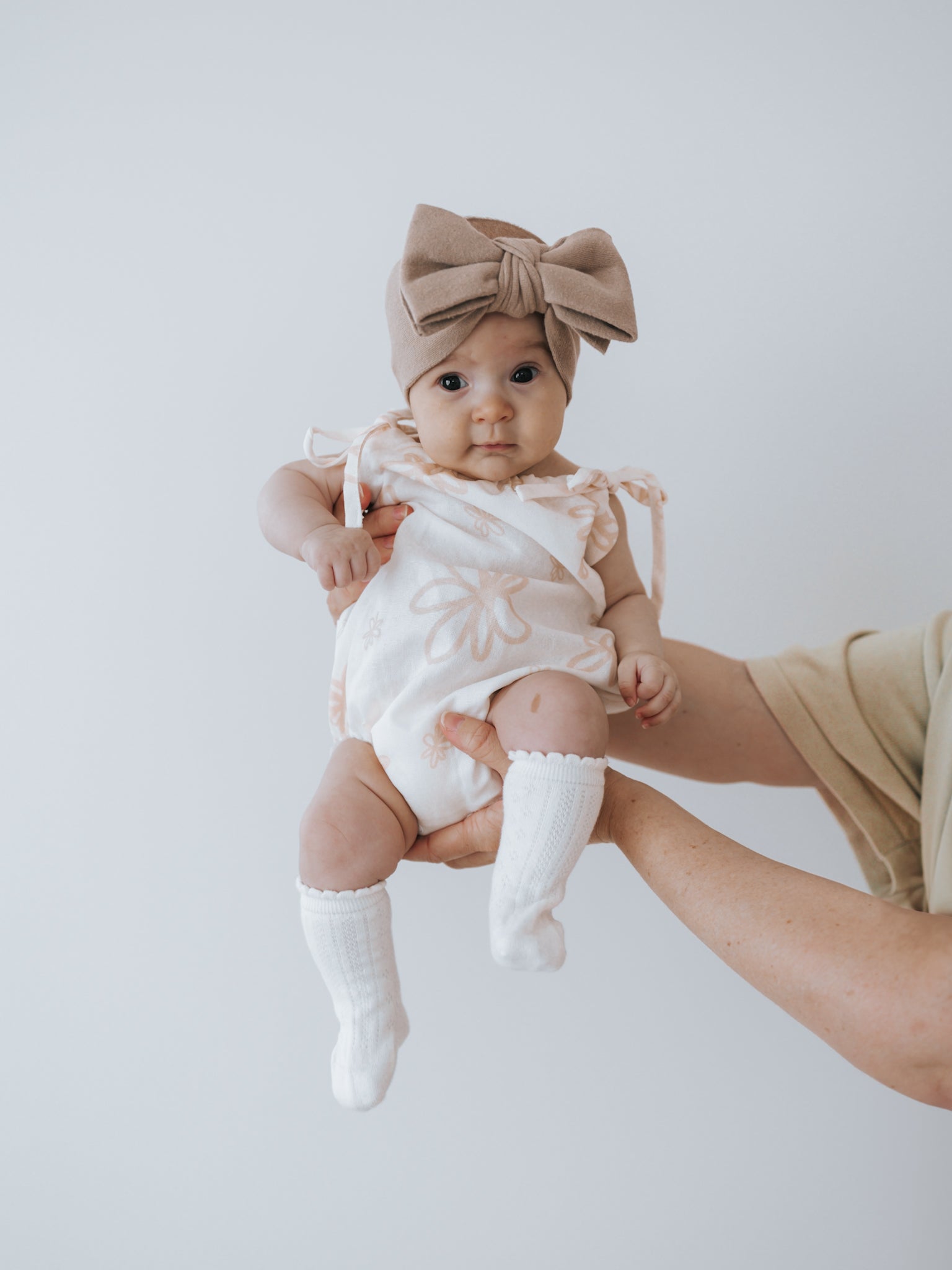 A baby, dressed in a delightful ZIGGY LOU Eva Bubble Romper Gia with a charming floral pattern, is paired with white socks and a large brown bow headband as they are held aloft against a plain backdrop.