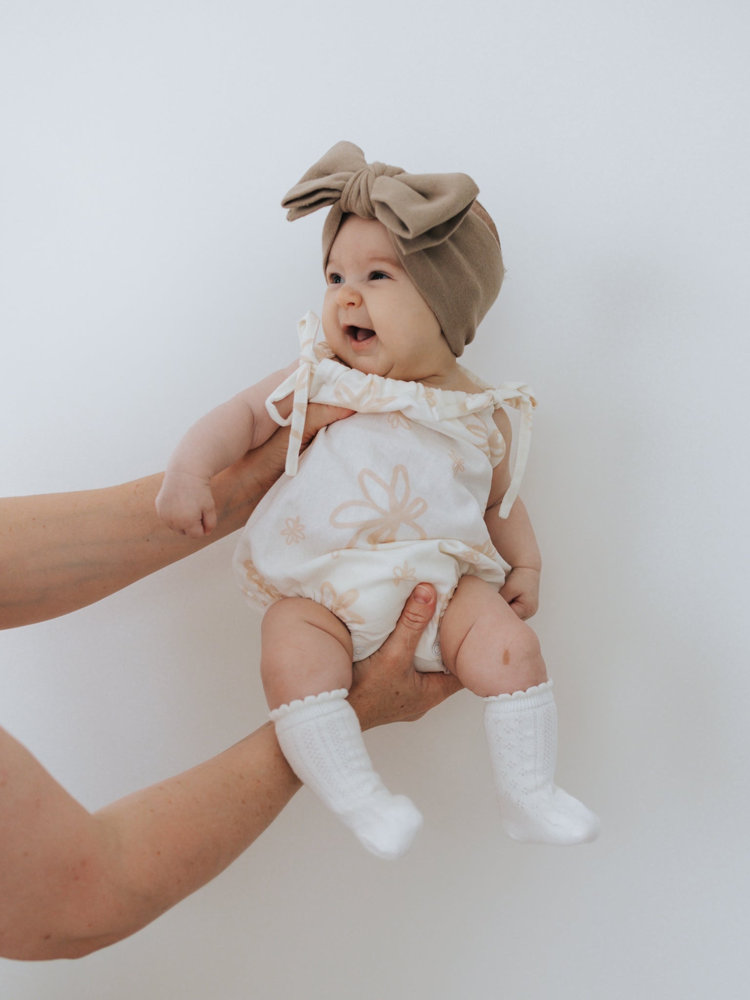 A smiling baby in a lightweight Eva Bubble Romper Gia by ZIGGY LOU, complemented by white socks and a large brown headband, is held up by two hands against a plain white background. The romper boasts a delicate Gia printed design, adding an extra touch of charm to the adorable outfit.