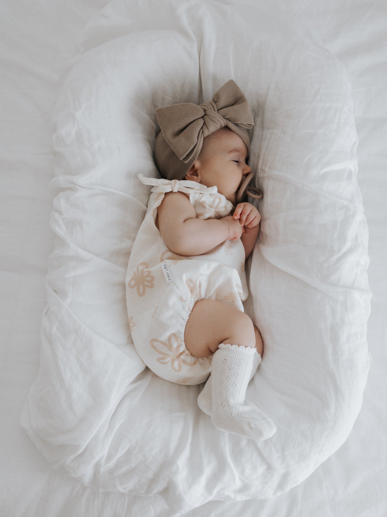 A baby wearing the Eva Bubble Romper Gia by ZIGGY LOU, complemented with a large brown bow headband and white socks, sleeps on a soft white cushion. The little one lies on their side with hands gently clasped near the face.