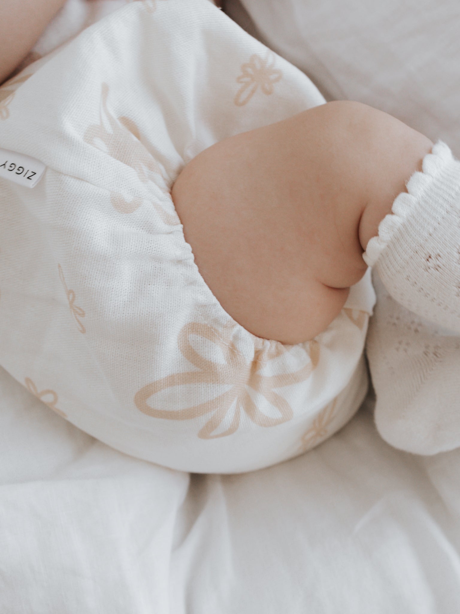 Close-up of a baby lying on a white bed, wearing the Eva Bubble Romper Gia from ZIGGY LOU, adorned with delicate floral patterns. The baby's small foot is wrapped in a white sock. The soft lighting creates a peaceful and gentle atmosphere.