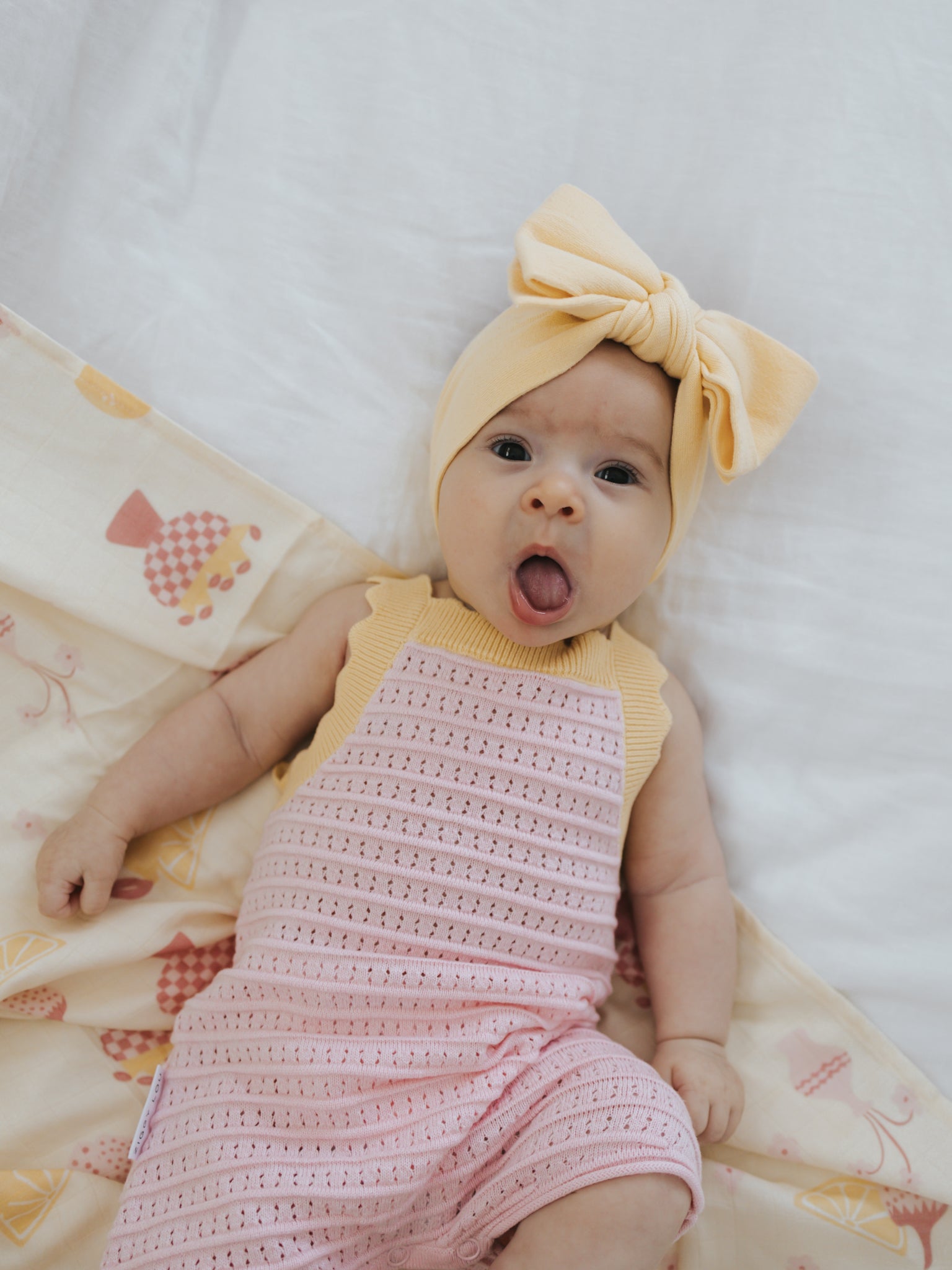 A baby dressed in the Playsuit Clementine by ZIGGY LOU, featuring a charming pink eyelet knit design and accessorized with a yellow headband adorned with a large bow, is lying on a patterned blanket. Crafted from 100% cotton, this adorable outfit complements the baby's open mouth and wide eyes, perfectly capturing an expression of surprise or curiosity.