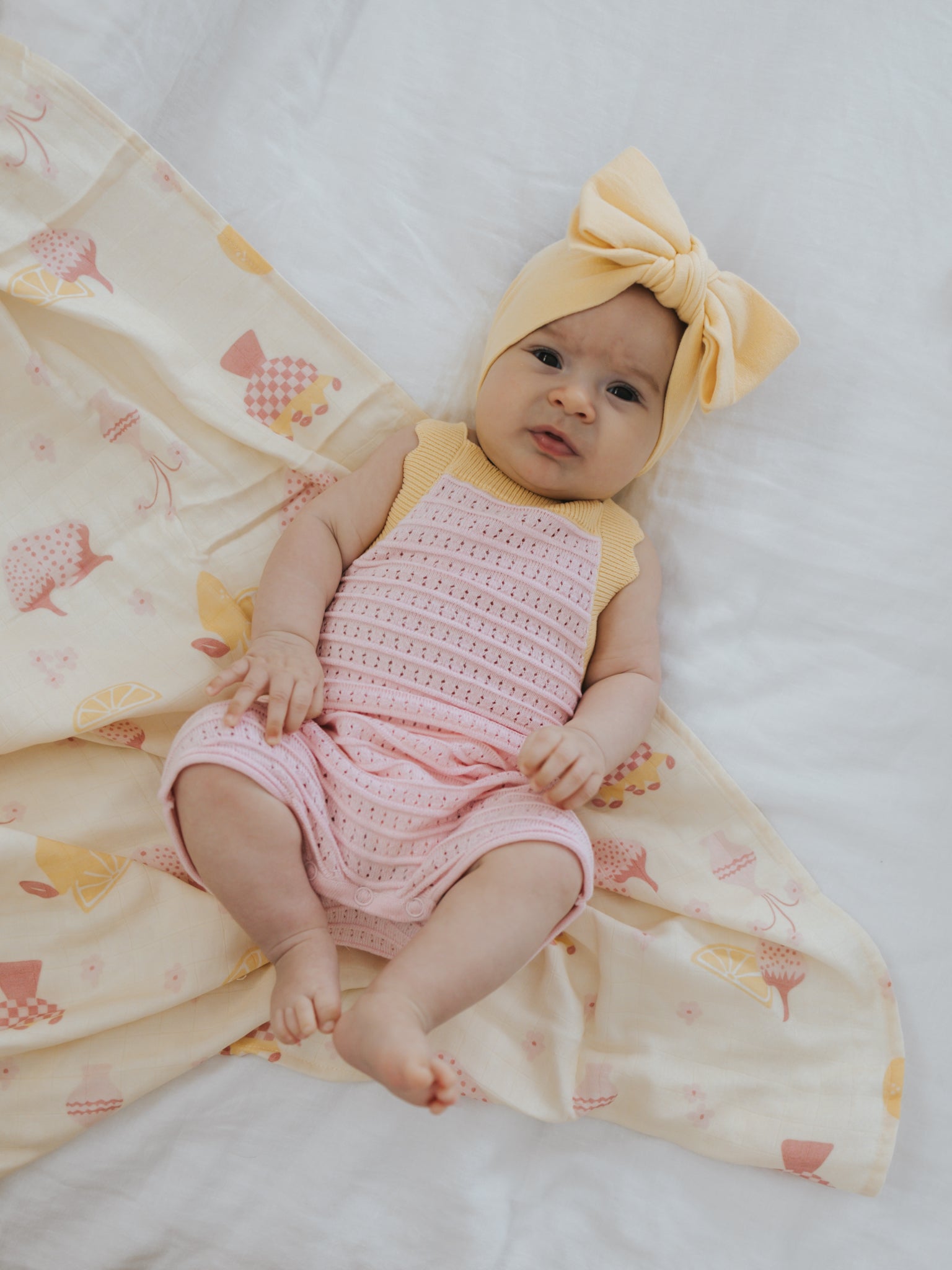 A baby rests on a white bed with a patterned blanket, snug in the Playsuit Clementine by ZIGGY LOU, crafted from 100% cotton in a charming pink eyelet knit. The large yellow headband with a bow adds charm, while the blanket displays playful chicks and umbrellas, complementing the calm expression on their face.