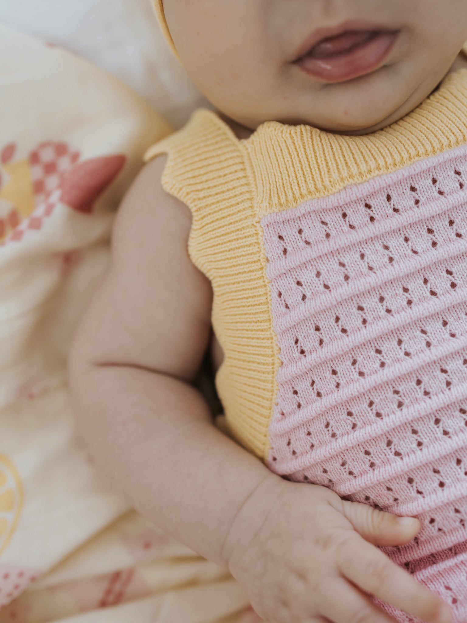 A baby in the charming ZIGGY LOU Playsuit Clementine, featuring pastel pink and yellow eyelet knit, rests on a patterned blanket. The close-up image focuses on the baby's torso, tiny hands, and mouth. Crafted from 100% cotton fabric, it lies on a blanket decorated with delicate floral and geometric designs.