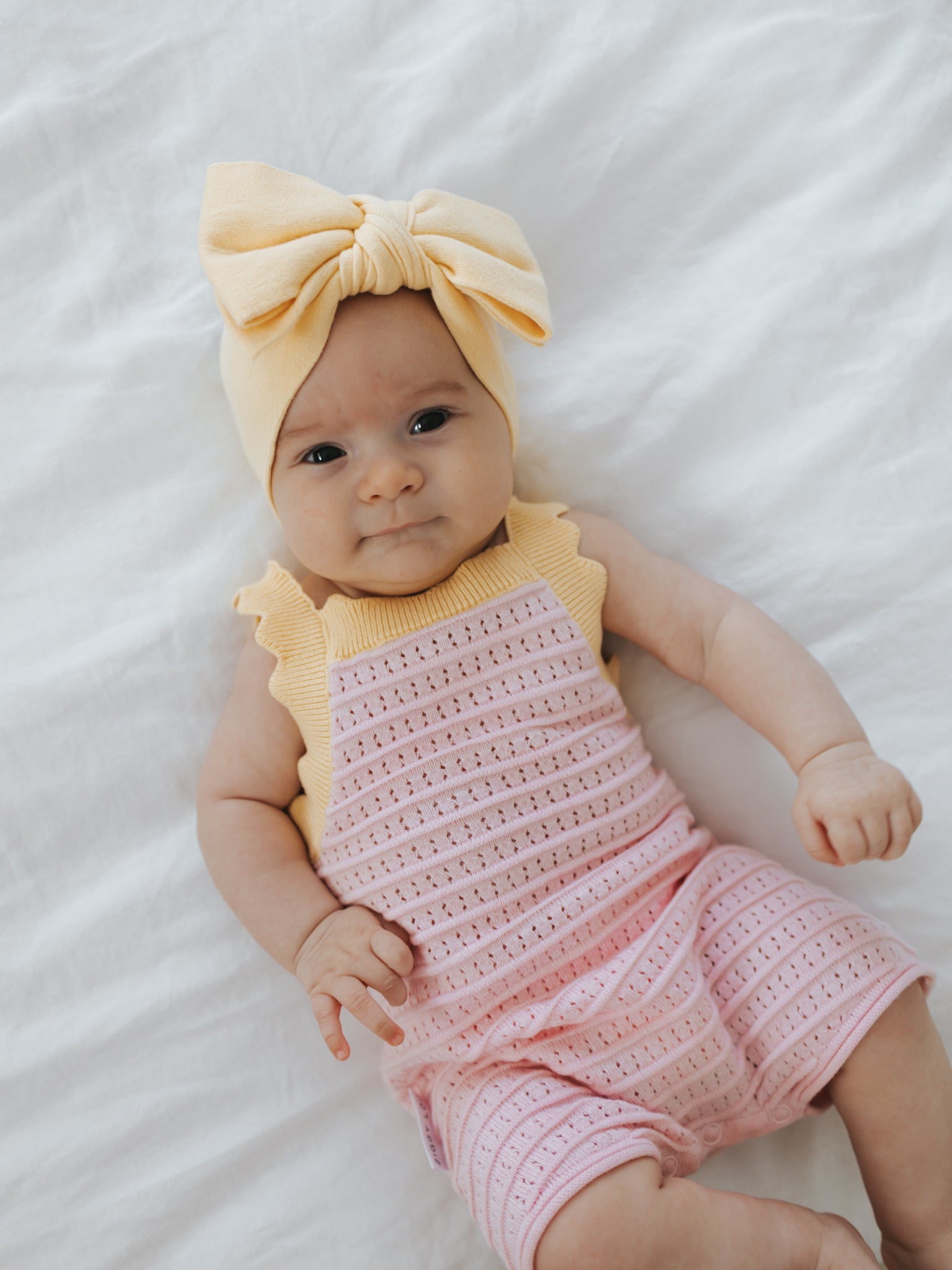 A baby wearing a yellow headband with a large bow and the Playsuit Clementine from ZIGGY LOU rests on a soft white blanket. The 100% cotton outfit makes the little one look content and relaxed.