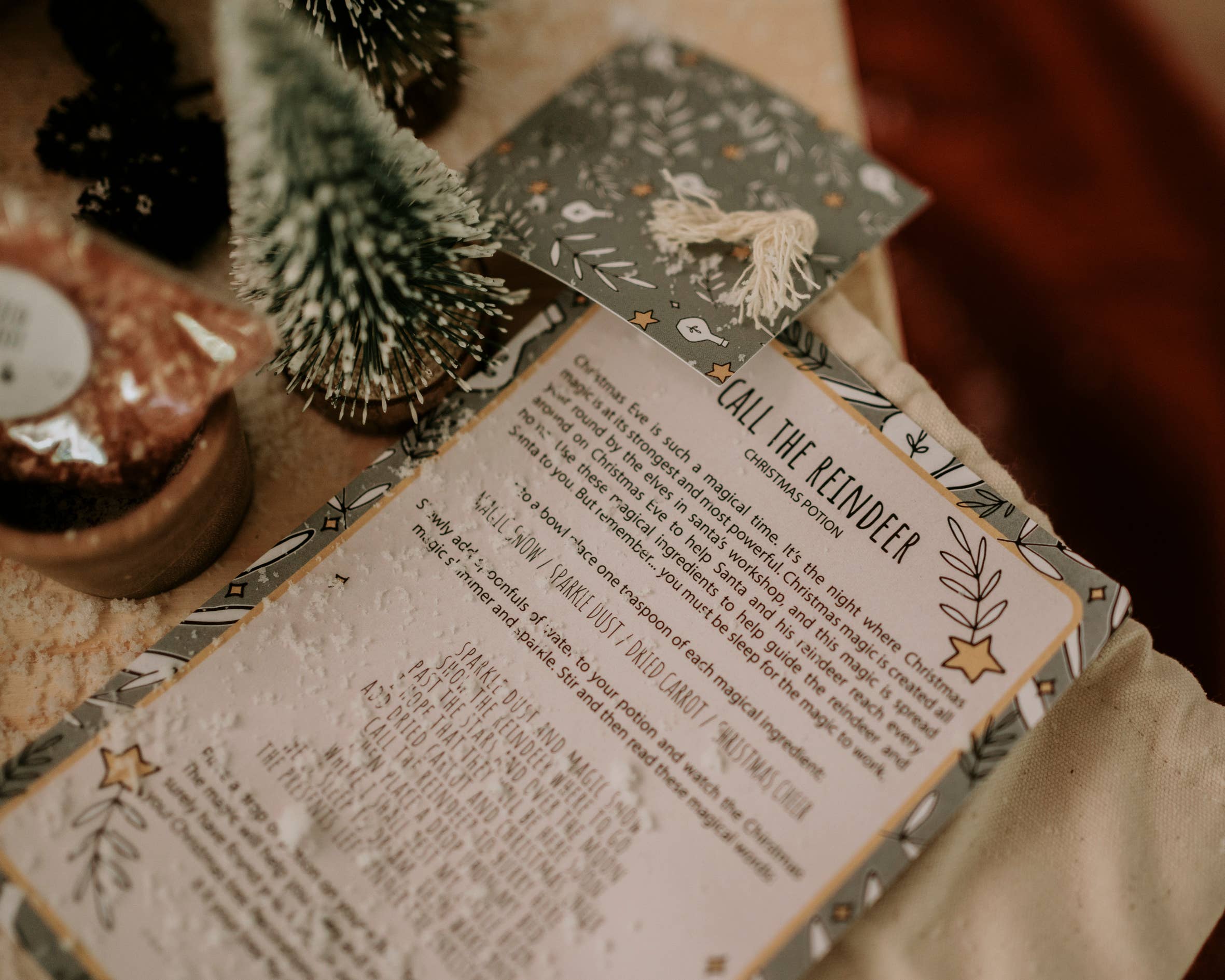 A festive pouch titled "Xmas Eve Potion Pouch to Call the Reindeer" from THE LITTLE POTION CO sits on a table, surrounded by miniature evergreen trees and holiday-themed paper. This magical tradition features decorative borders with stars and foliage, inviting enchantment into the holiday season.