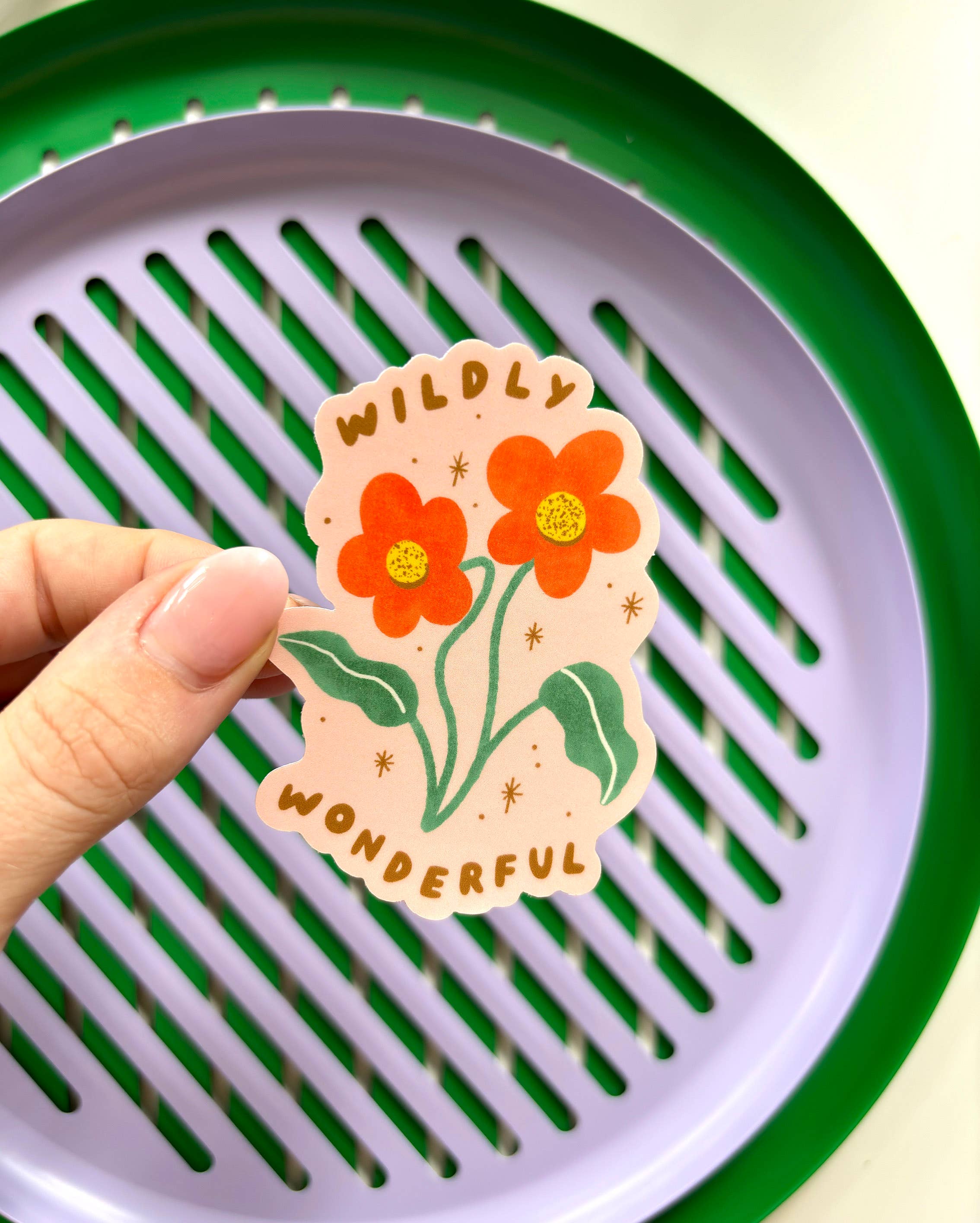 A hand holds a "Wildly Wonderful Sticker" from LAUREN SISSONS STUDIO, showcasing two red-orange wildflowers and the words "Wildly Wonderful." The background consists of a lilac-colored tray with green edges featuring slotted grooves. The overall tone is cheerful and vibrant, making this vinyl delight stand out beautifully.