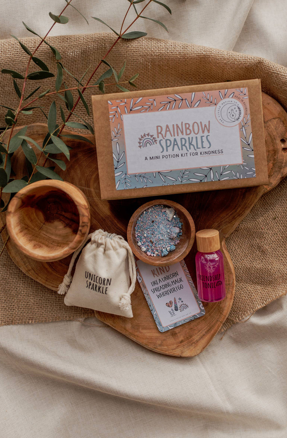 A flat lay of the MINI Rainbow Sparkles Potion Kit (A potion for kindness) from THE LITTLE POTION CO on a wooden tray. Perfect for imaginative play, the kit includes a small bottle labeled "Friendship Tonic," a cloth bag labeled "Unicorn Sparkle," a packet of glitter, and an instruction card. Eucalyptus leaves adorn the scene.