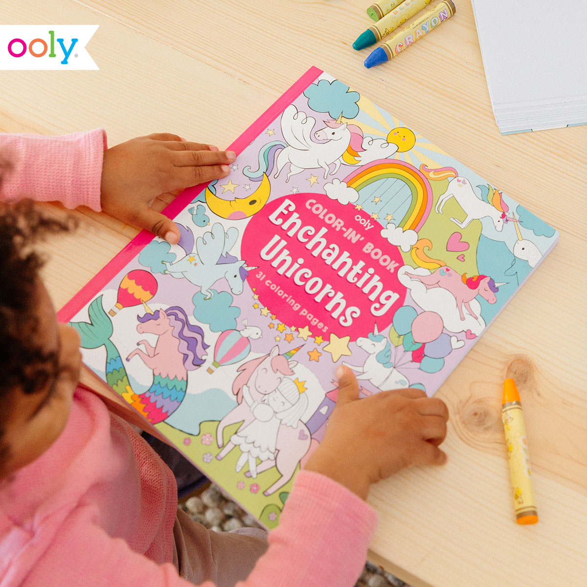 a young girl with the OOLY colour in book "enchanting unicorns" on a table with crayons surrounding it.