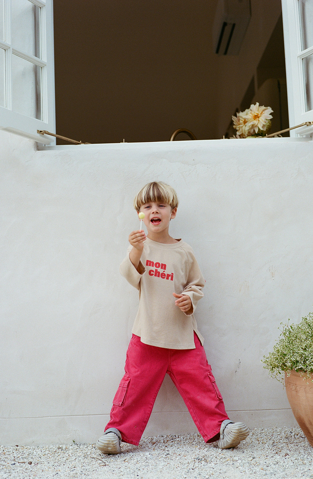 A young child with a blond bowl haircut stands outdoors against a white wall. The child wears a beige "mon chéri" shirt and bright red Louie Denim Jeans from THE WHOLESOME STORE, holding something small and white in their right hand. Nearby, a plant with tiny white flowers is partially visible.