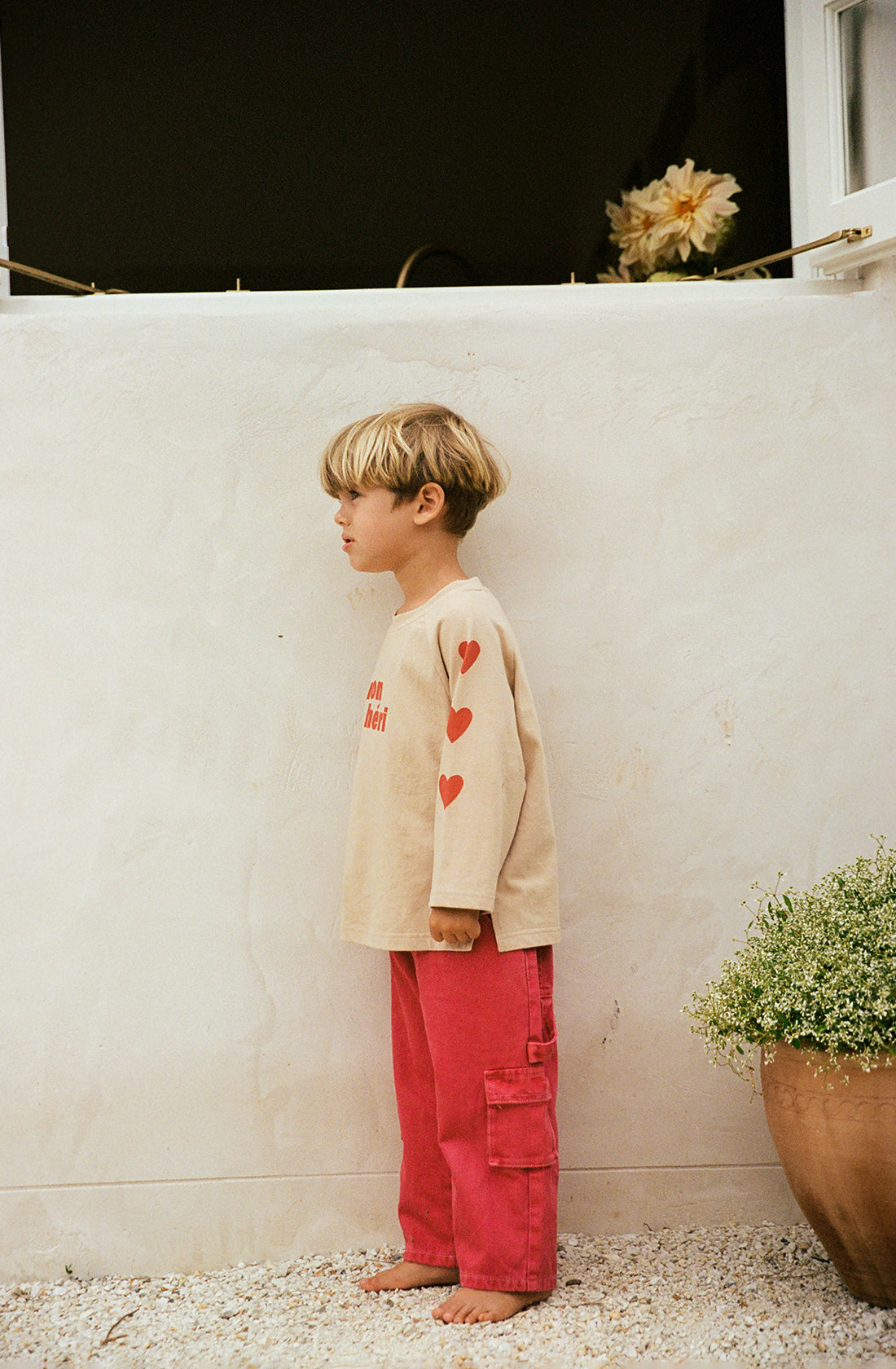 A young child stands barefoot on a gravel surface, wearing a beige long-sleeve shirt with heart designs on the sleeves and Louie Denim Jeans Vintage Red from THE WHOLESOME STORE. They are standing next to a white wall with a potted plant nearby. The background includes part of a building and some flowers.