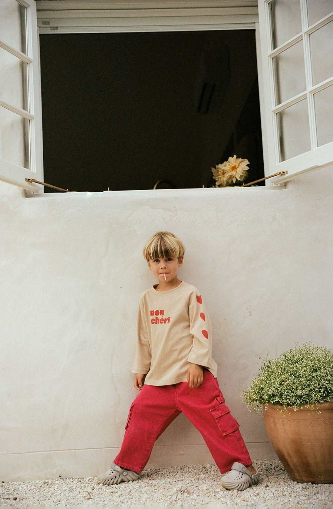 A young child with a bowl haircut stands outdoors against a white wall under an open window. The child wears a beige shirt with red text and THE WHOLESOME STORE's Louie Denim Jeans Vintage Red featuring an elastic waistband. A potted plant is visible on the right side, adding to the bright and casual setting.