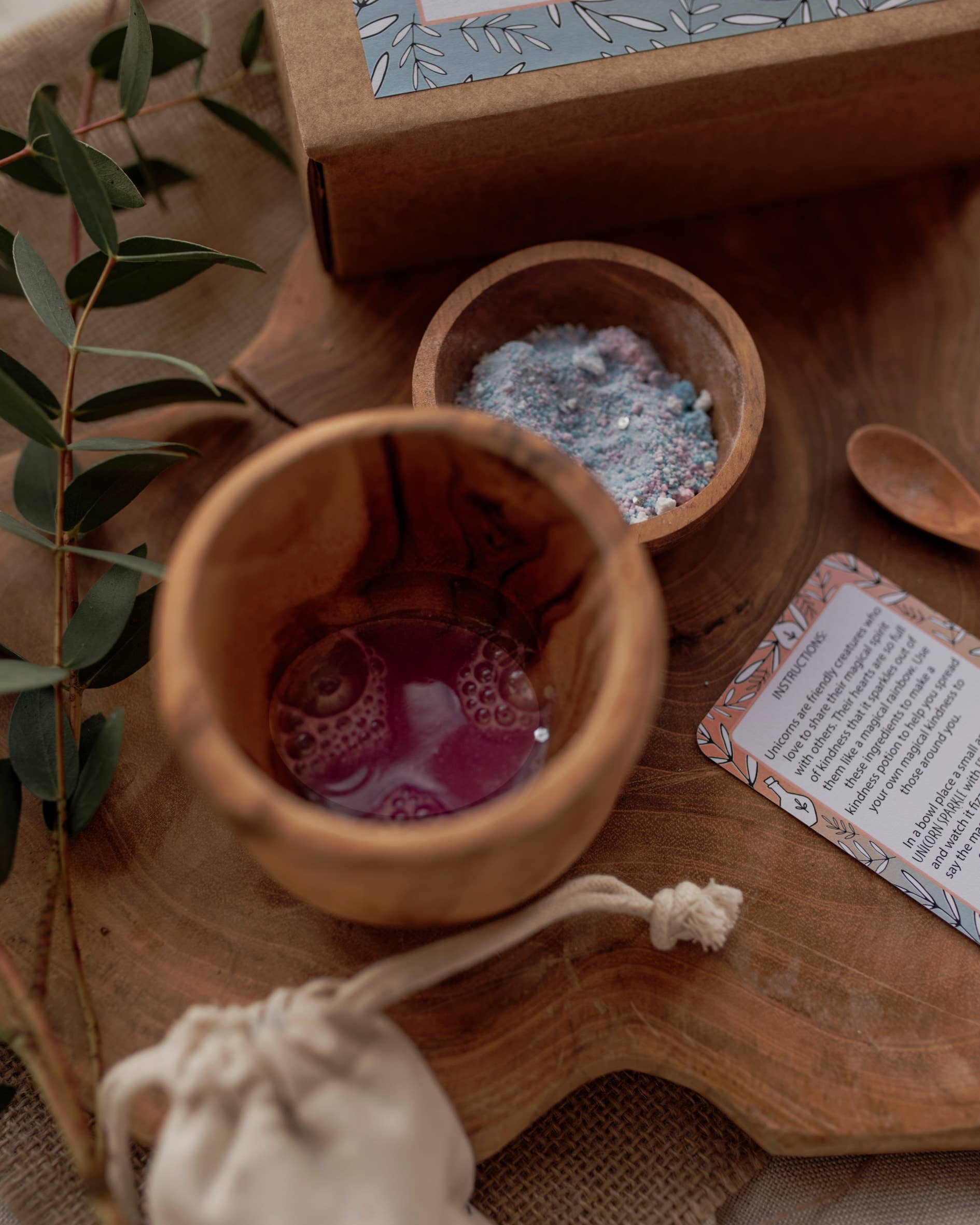 The MINI Rainbow Sparkles Potion Kit (A potion for kindness) by THE LITTLE POTION CO features a wooden cup with a bubbly liquid, a wooden bowl filled with colorful powder, and a small spoon, all arranged on a wooden surface with green leaves. Also included are a cloth bag and card. In the background, the enchanting mindfulness kit is completed by a floral design box.