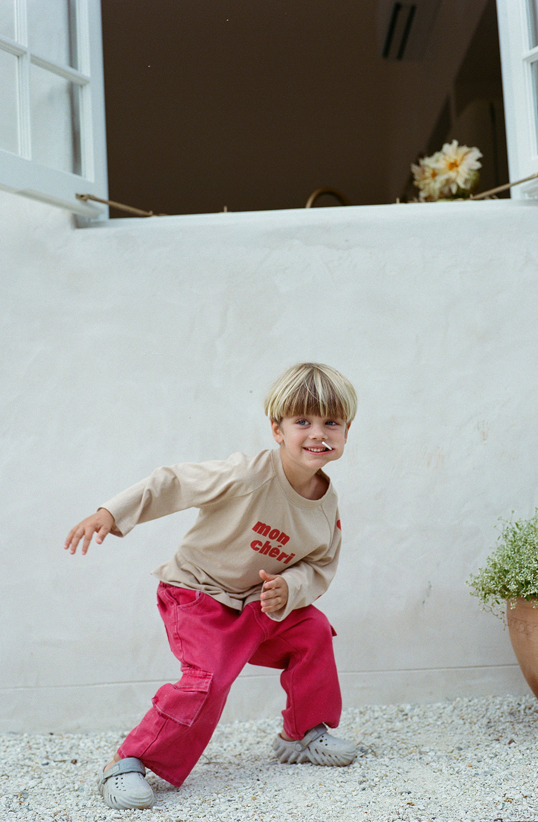 A young child with blonde hair is smiling and striking a playful pose while wearing a beige shirt with red letters and a pair of bright pink, wide-leg pants called Louie Denim Jeans Vintage Red by THE WHOLESOME STORE. The child stands on a white gravel surface in front of a white wall with two open windows, and there’s a potted plant visible on the right.