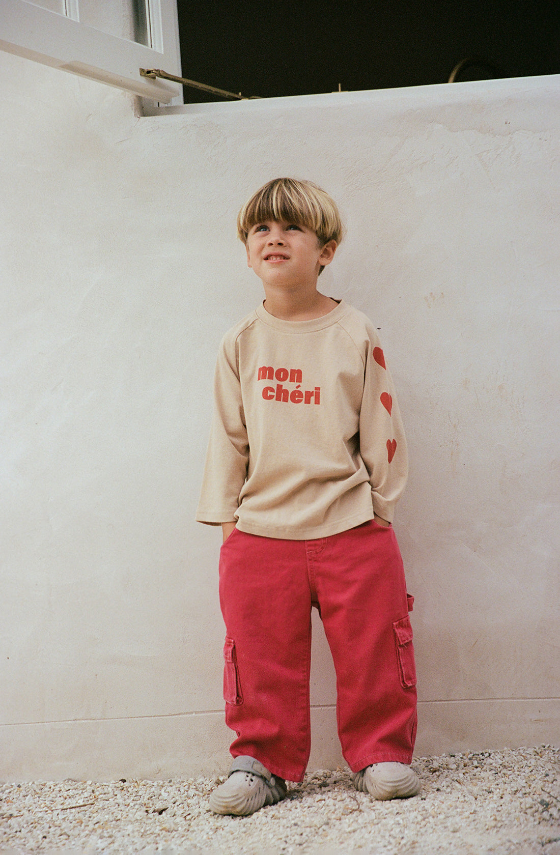 A child stands outdoors against a white wall, wearing a beige long-sleeve shirt with "mon chéri" and red hearts on the sleeves, paired with THE WHOLESOME STORE's Louie Denim Jeans in Vintage Red, and white shoes. The child has short blonde hair and stands with hands behind their back, smiling slightly.