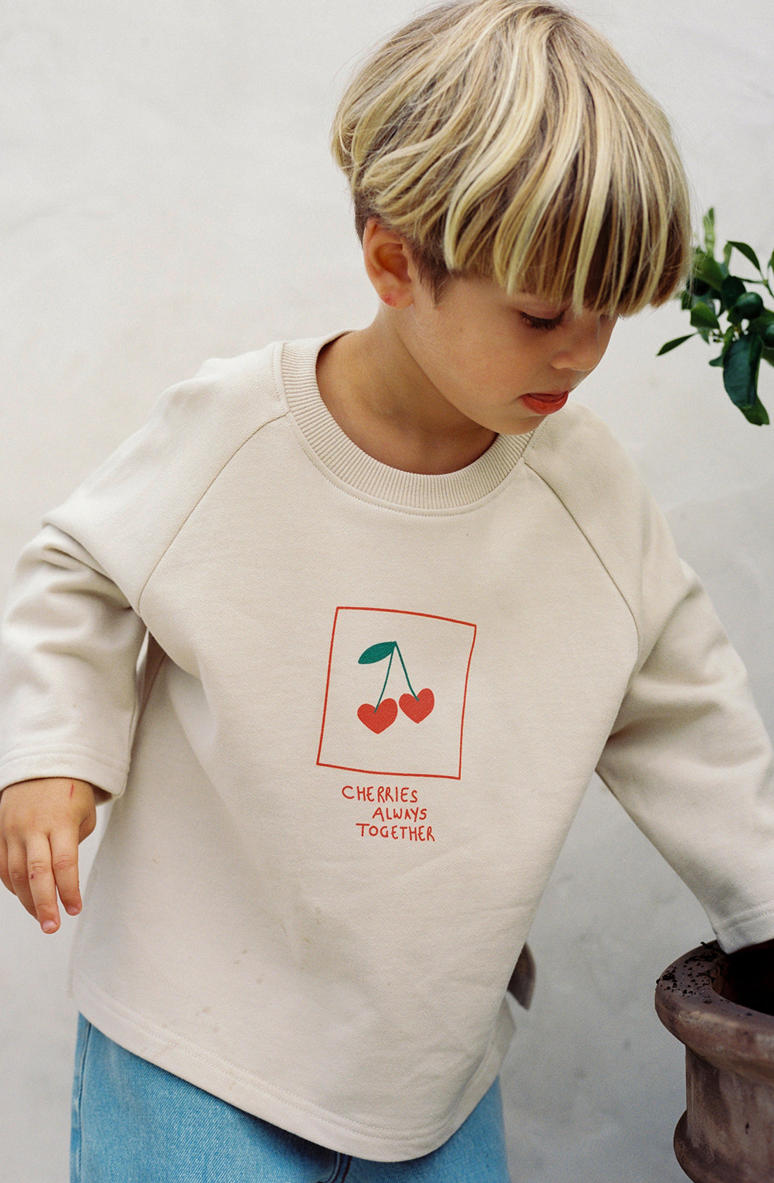 A young child with light blonde hair is wearing the Cherries Pull Over from THE WHOLESOME STORE. This cream-colored sweatshirt, crafted from 100% cotton, showcases an illustration of two cherries alongside the text "CHERRIES ALWAYS TOGETHER." The child gazes downward and slightly to the side, with greenery faintly visible in the background.