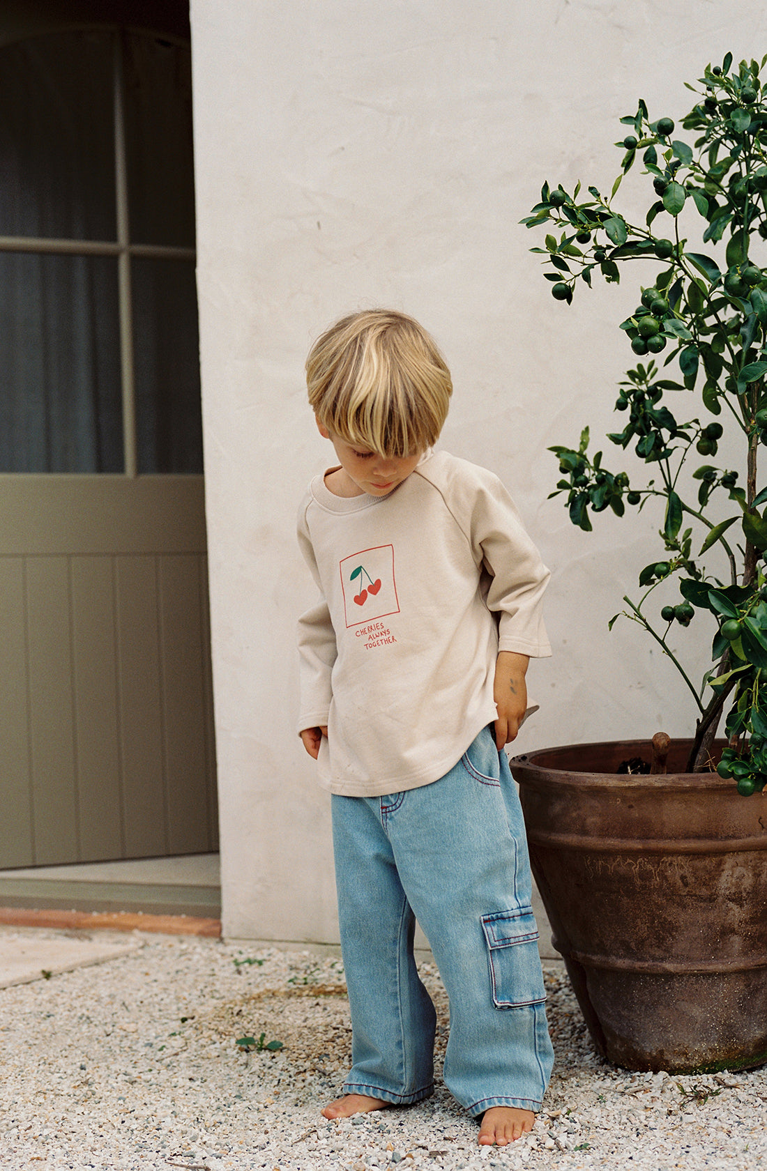 A young child with blond hair stands barefoot on a gravel path in front of a white wall. The child is wearing THE WHOLESOME STORE's ethically made Cherries Pull Over and light blue jeans with loose-fitting cargo pockets. Next to the child is a large potted plant.