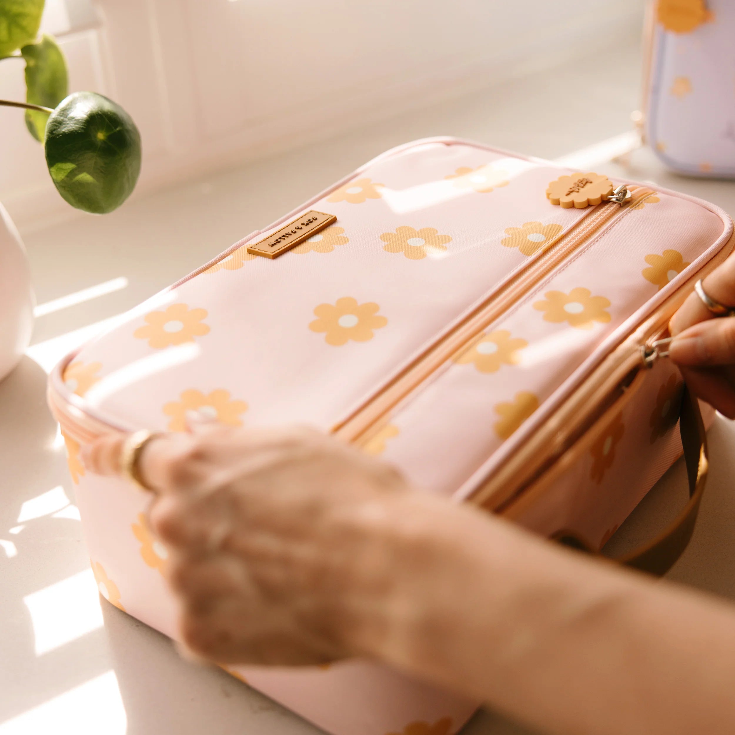 A person is zipping up a FOX & FALLOW Daisy Chain Lunch Bag, featuring a peach color and yellow flower patterns that are easy to clean. The insulated lunch bag rests on a white surface near a sunlit window, with green leaves visible in the background.