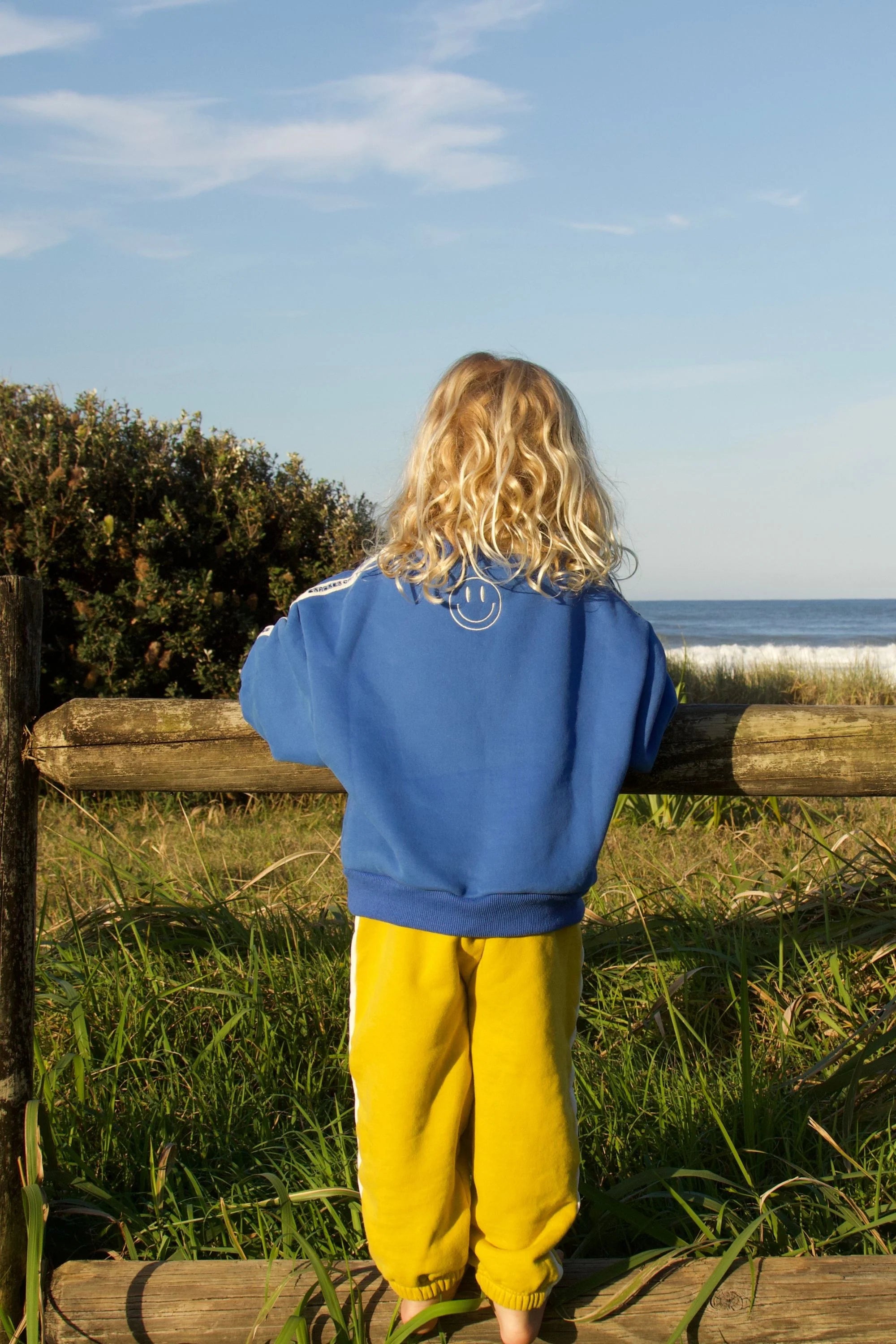 A young child with long blond hair, wearing the CRACKER DAY CLUB Cracker Day Club - Kids Cloud Racer Sweater Blue and yellow pants, stands on grass facing away, looking at the ocean over a wooden fence.