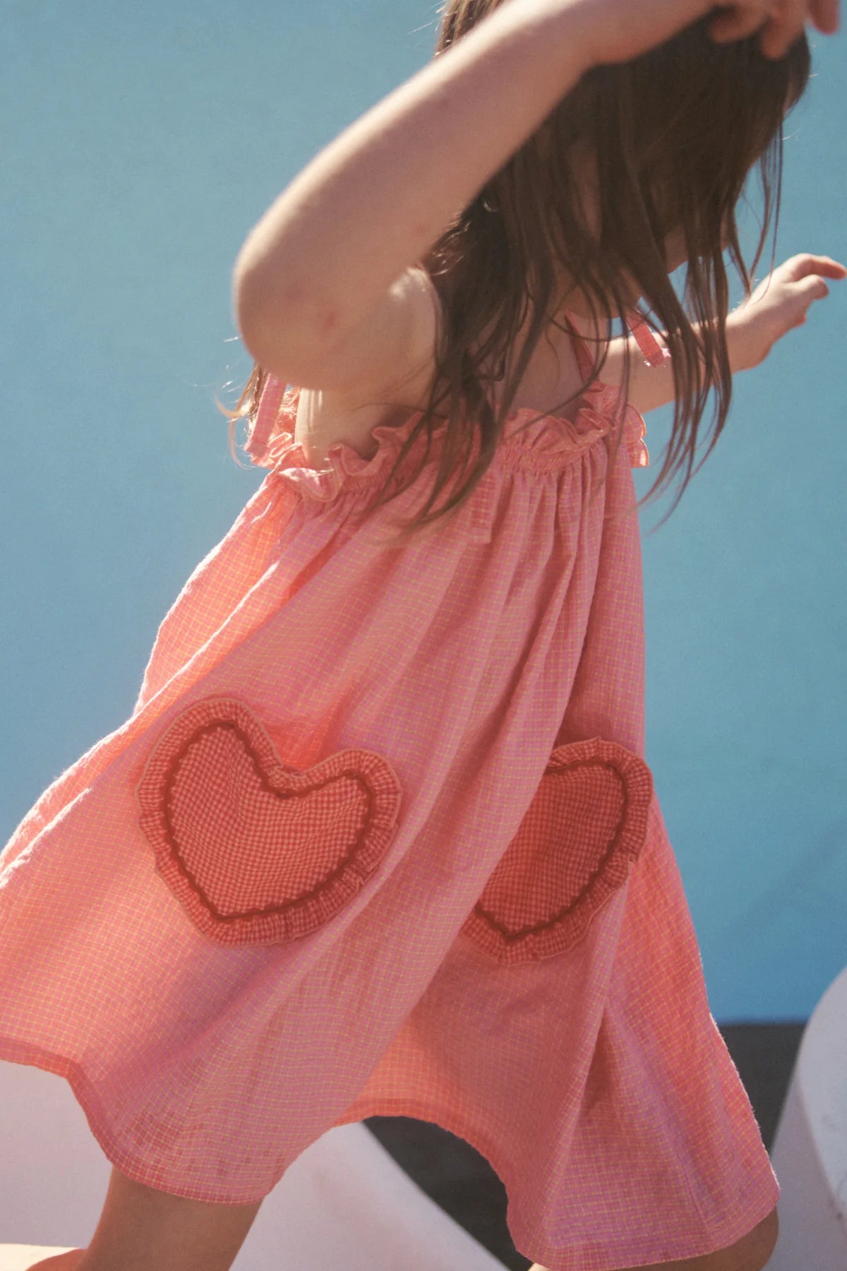 A young girl joyfully plays outdoors in the Lola Dress Pink Lemonade by LITTLE THE LABEL. The cotton dress features heart-shaped pockets, set against a sunny backdrop with a blue sky. Her arms spread wide as she delights in the moment.