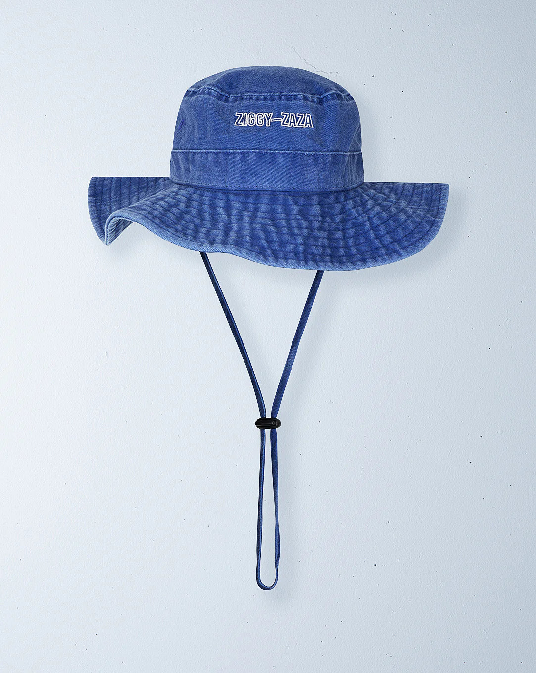 A blue denim Everyday Scout Hat Blu by ZIGGY ZAZA hangs on the wall, featuring an adjustable chin strap, embroidered "Ziggy Zaza" text on the front, and cotton canvas stitching details for a casual flair.