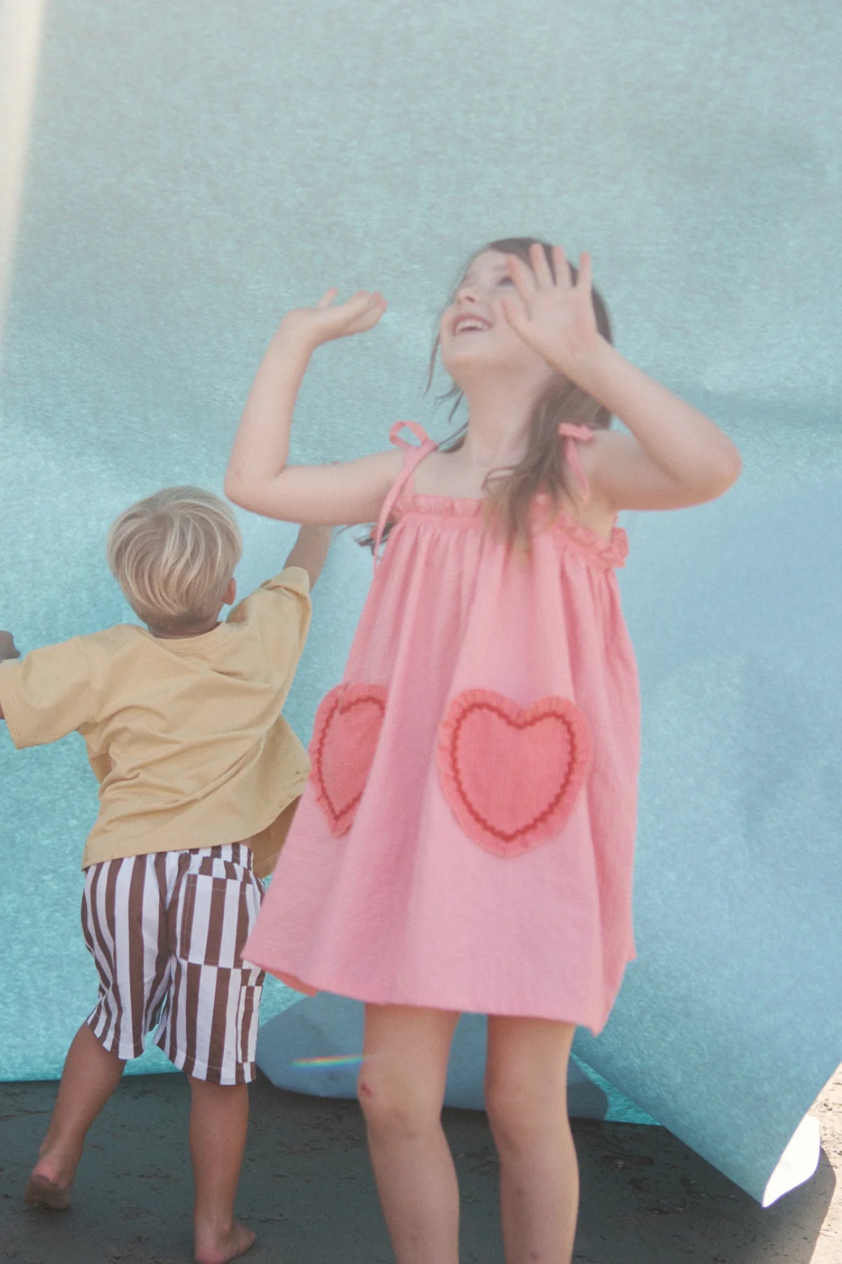 Two children play under a large blue sheet like a canopy. The girl, in LITTLE THE LABEL's pink Lola Dress with heart pockets, raises her arms joyfully. The boy, in a yellow shirt and striped shorts, is turned away as he enjoys the play.