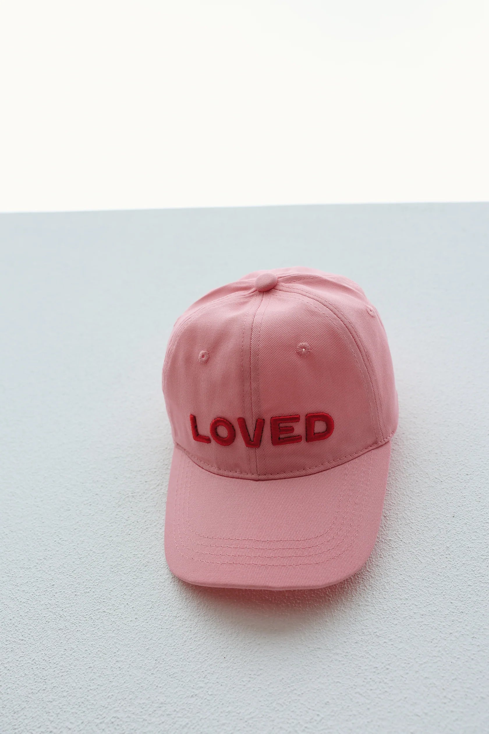 The TINY LOVE CLUB Loved Hat - Pink, featuring "LOVED" embroidered in red on the front, is displayed against a light textured background. This dad style cap has a six-panel design and an adjustable back strap, making it ideal for little heads.