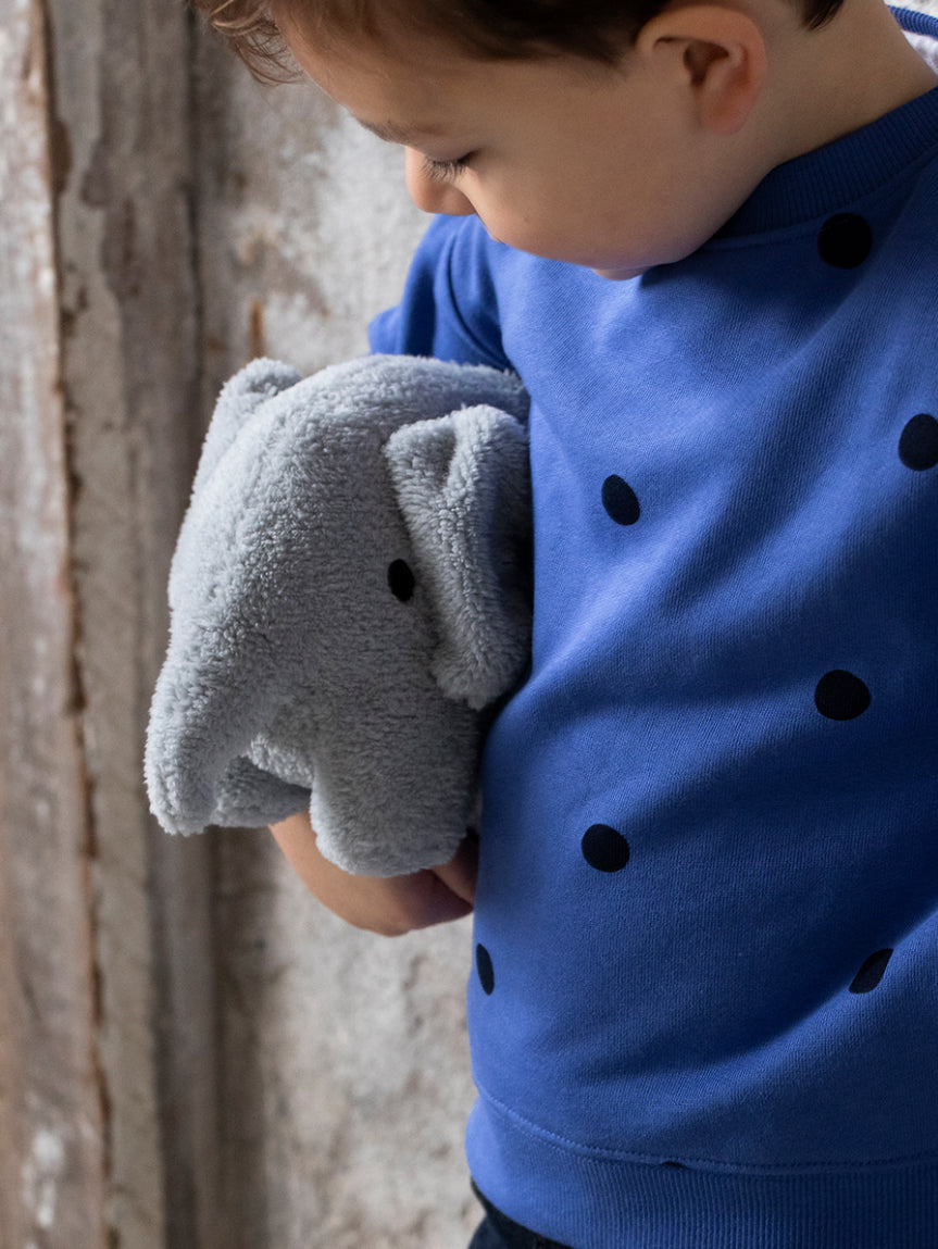 A young child in a blue sweater with black polka dots holds their BON TON TOYS X MIFFY Elephant Terry Light Grey 23 cm soft toy close to their chest. The child is looking down at the toy, which features a recycled PET filling. The background is a rustic wooden wall.