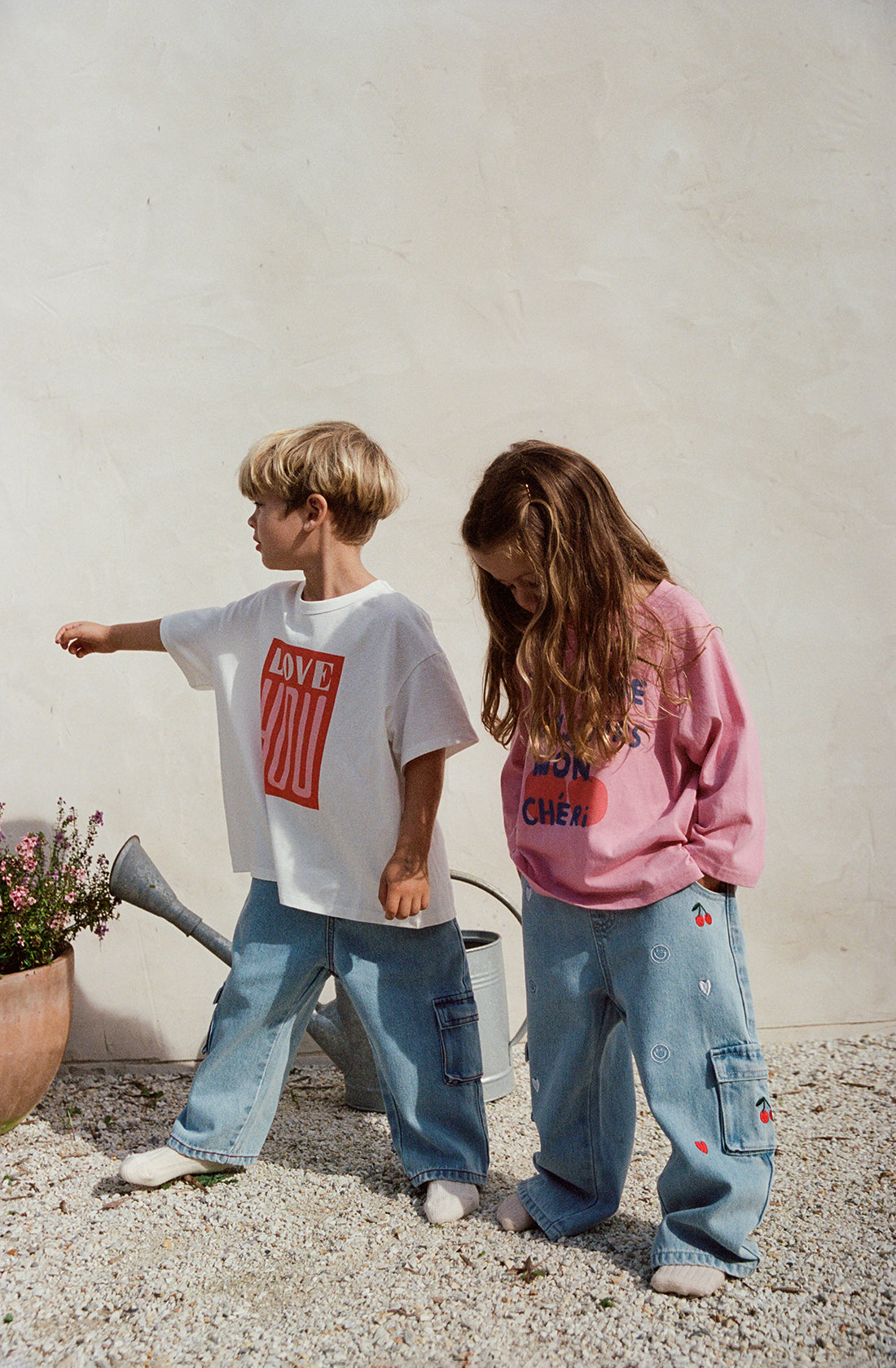 Two children standing outdoors against a white wall. One child is wearing a white T-shirt with "LOVE" written on it and THE WHOLESOME STORE's Louie Denim Jeans Mon Cheri, pointing towards a potted plant. The other child, in a pink sweatshirt and wide leg jeans, looks down at the ground.