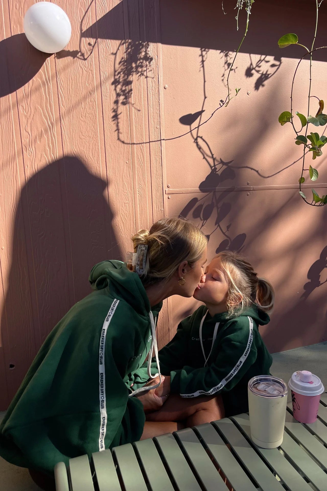 A woman and a young girl in CRACKER DAY CLUB Cloud Racer Hoodies sit together outdoors, sharing a kiss by a soft pink wall with plant shadows. On the table in front of them are drinks. The girl wears the Kids Cloud Racer Hoodie Forest.